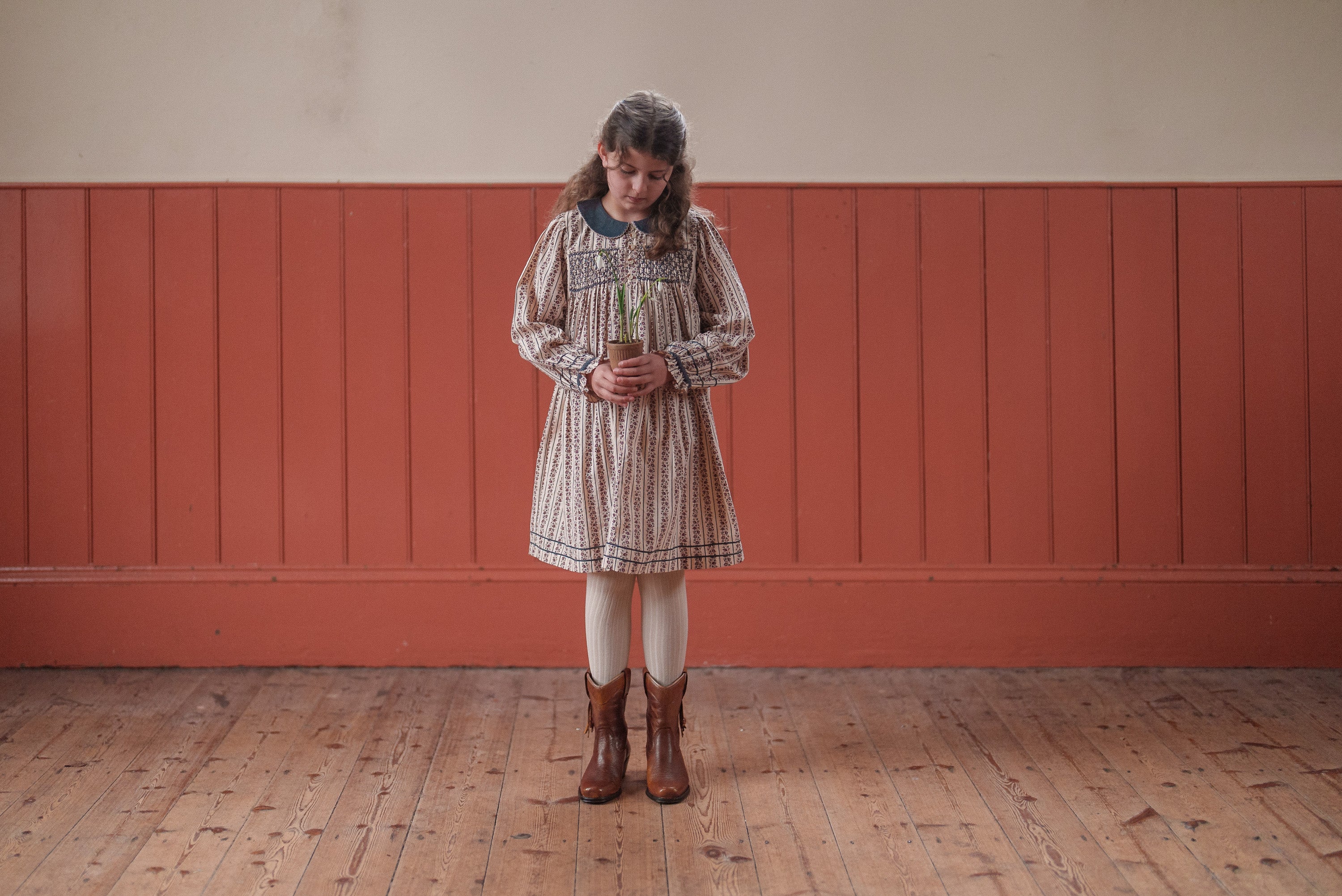 Young girl wearing a vintage-style dress made from organic cotton, standing on wooden floor against a rustic red paneled wall, embodying natural and ethical children's clothing.