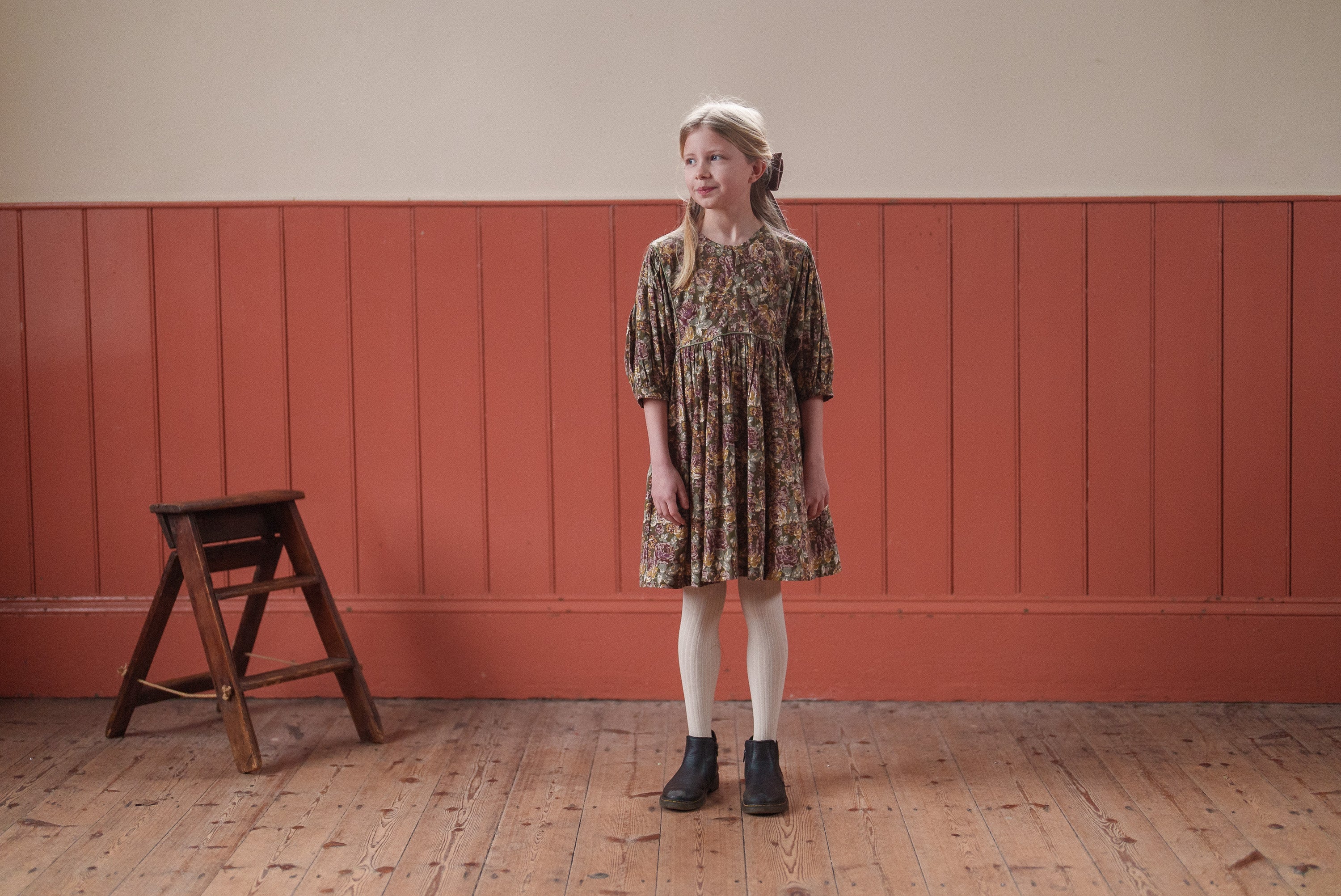 Young girl wearing a vintage style dress with a green floral pattern made from organic cotton, standing on a wooden floor against a warm-toned wall, showcasing sustainable kids clothing.