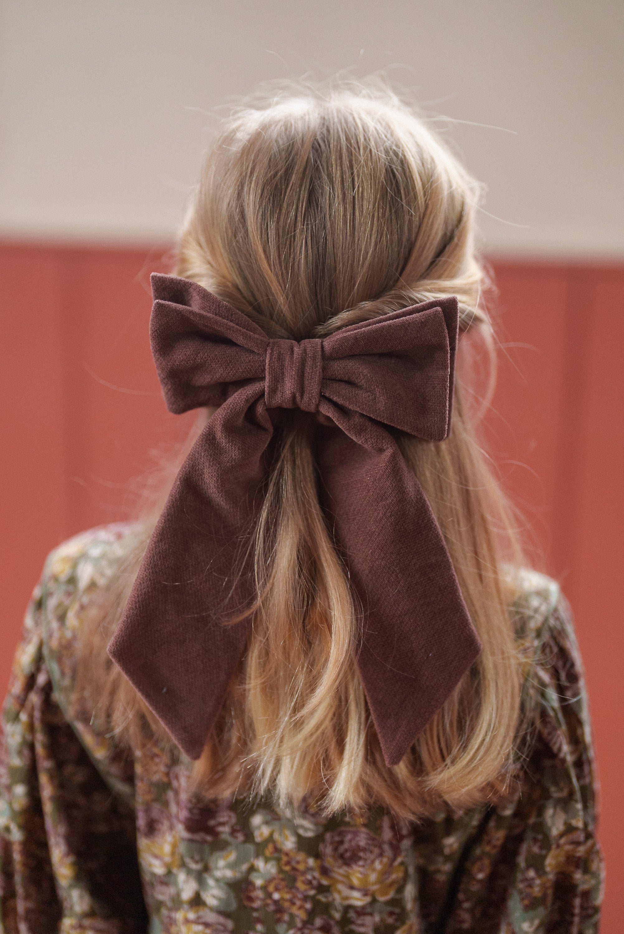 Young girl with blonde hair styled with a large brown fabric bow, wearing a vintage-style floral dress made from soft organic cotton.