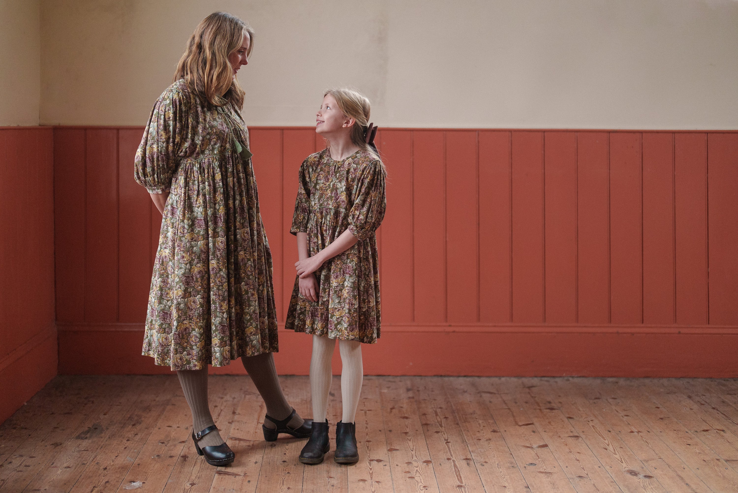 Mother and daughter wearing vintage style dresses made from organic cotton with a floral green pattern, showcasing ethically made, sustainable kids clothing in a warm, natural setting.