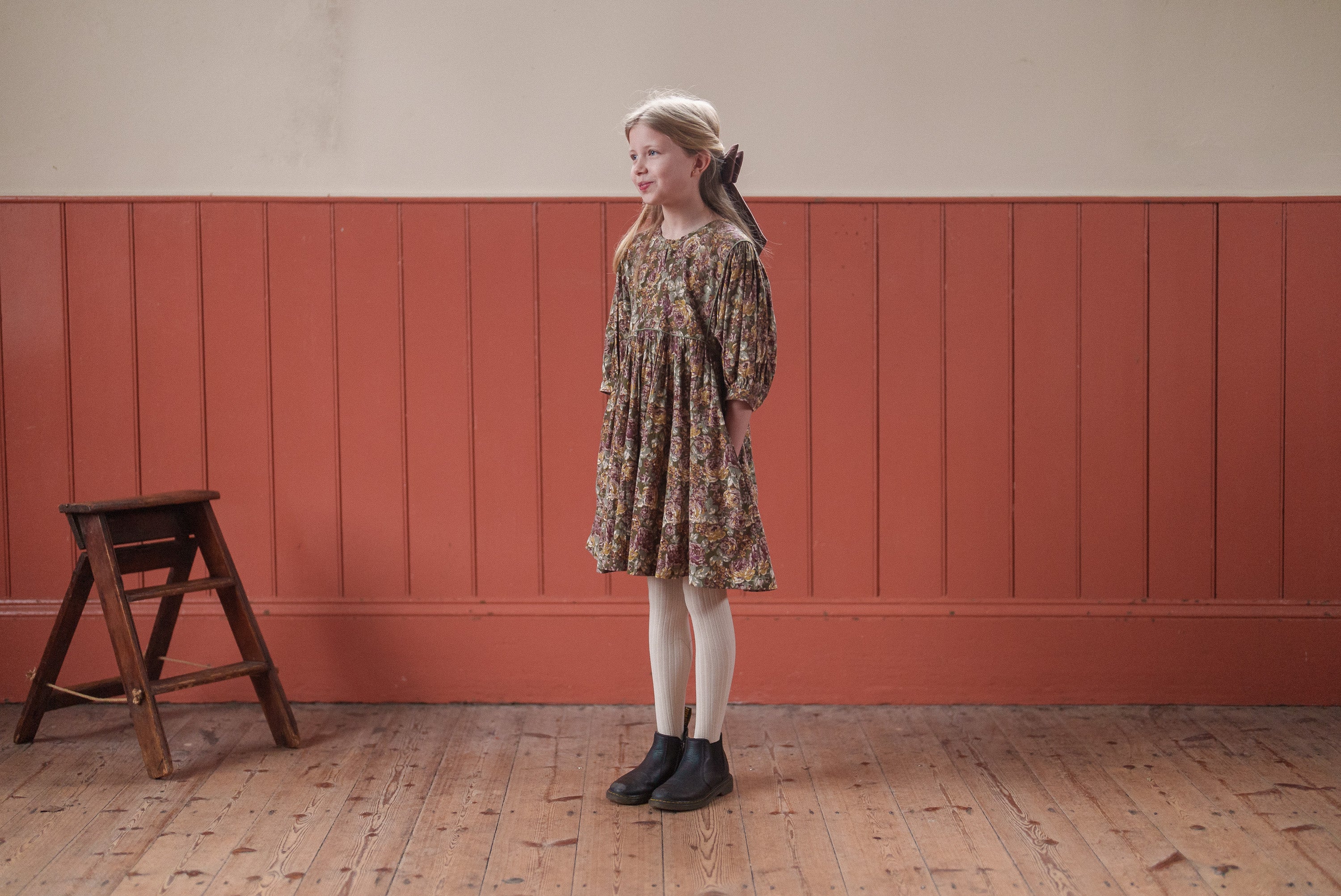 Young girl wearing a vintage style organic cotton floral dress in green tones, standing on a wooden floor next to a small wooden stool in a warm, natural setting.