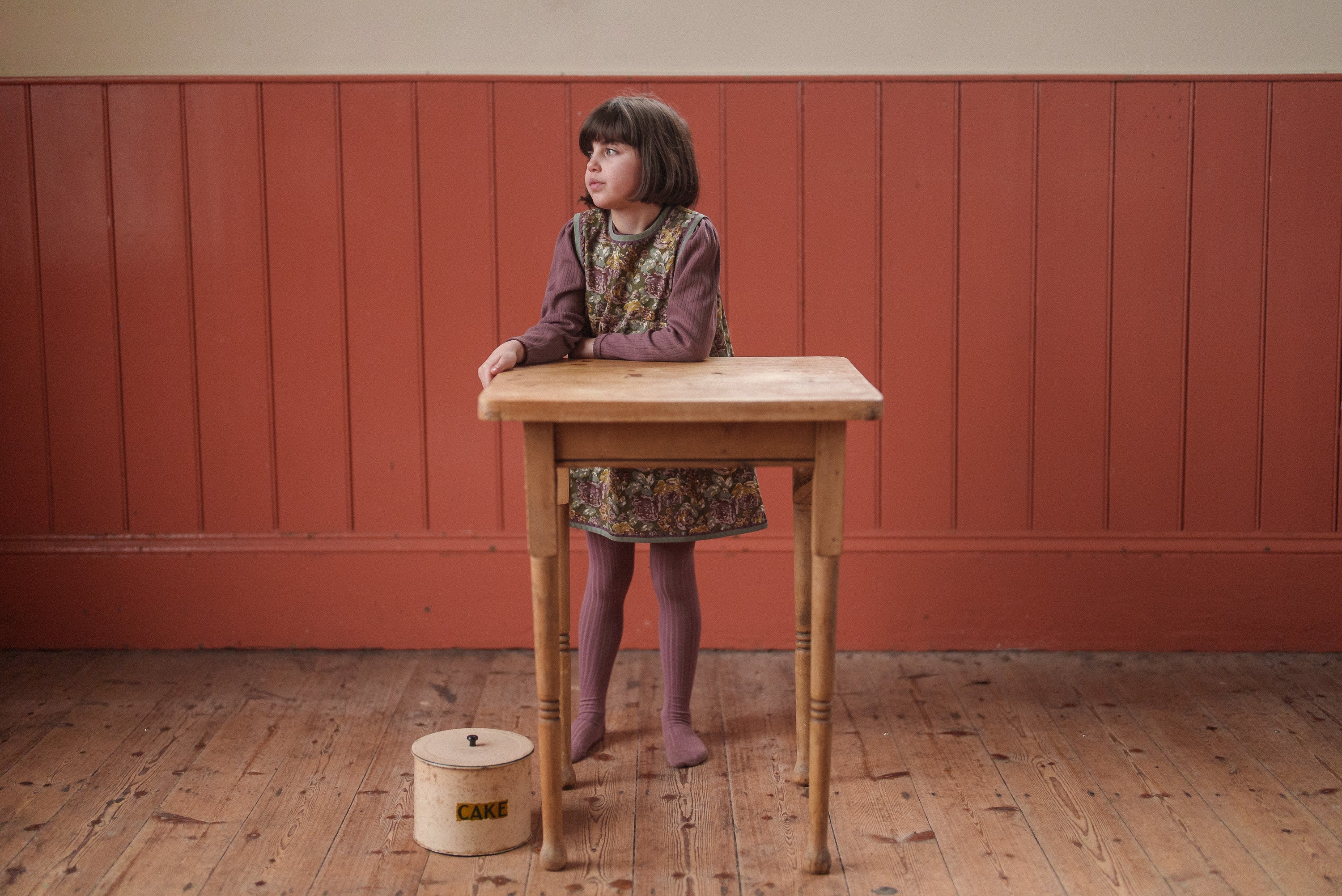 Young girl wearing a green floral organic cotton dress with vintage style standing behind a wooden table in a warm, rustic room.