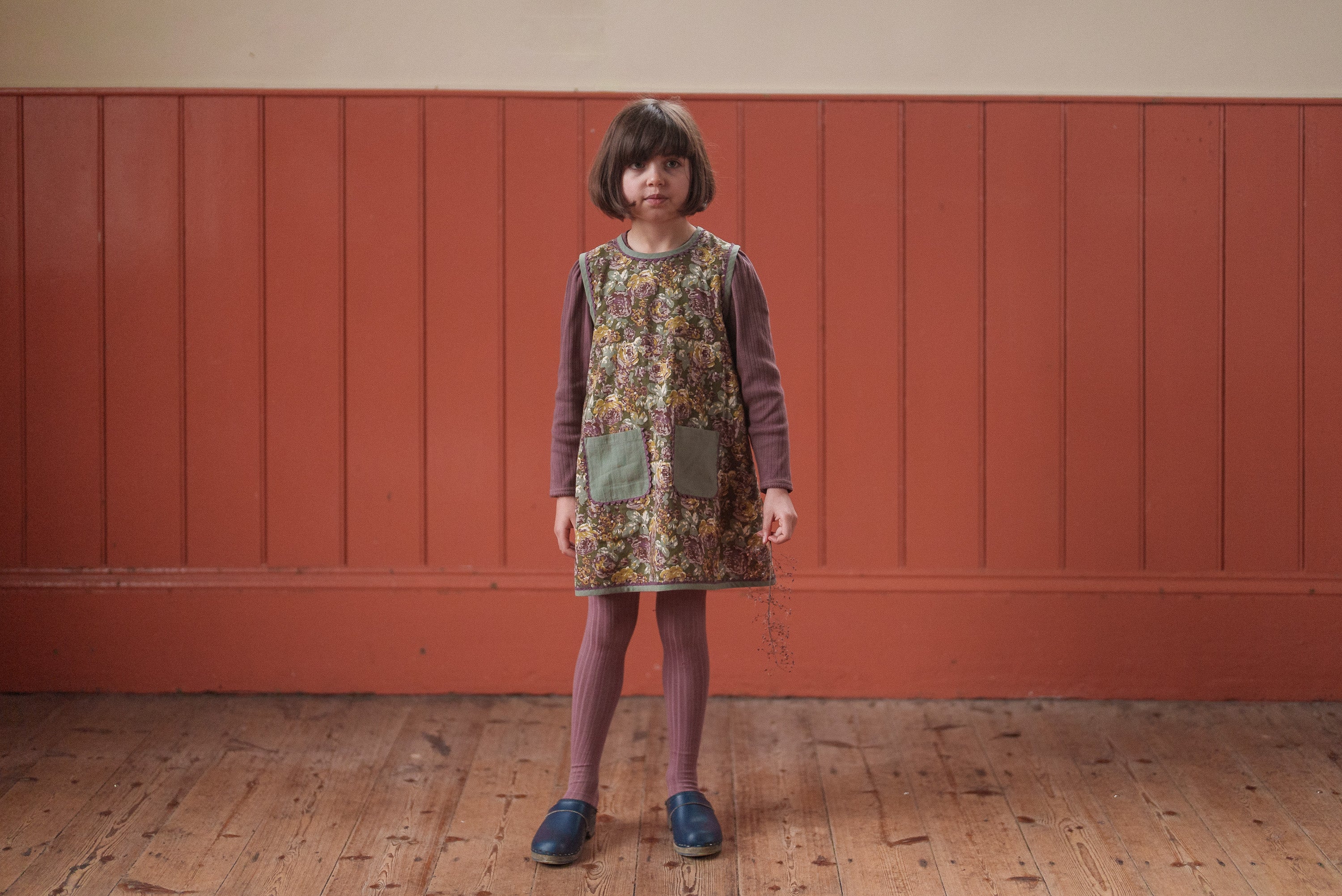 Young girl wearing a vintage style green floral dress made from organic cotton, paired with matching tights and comfortable blue shoes, standing on a wooden floor.