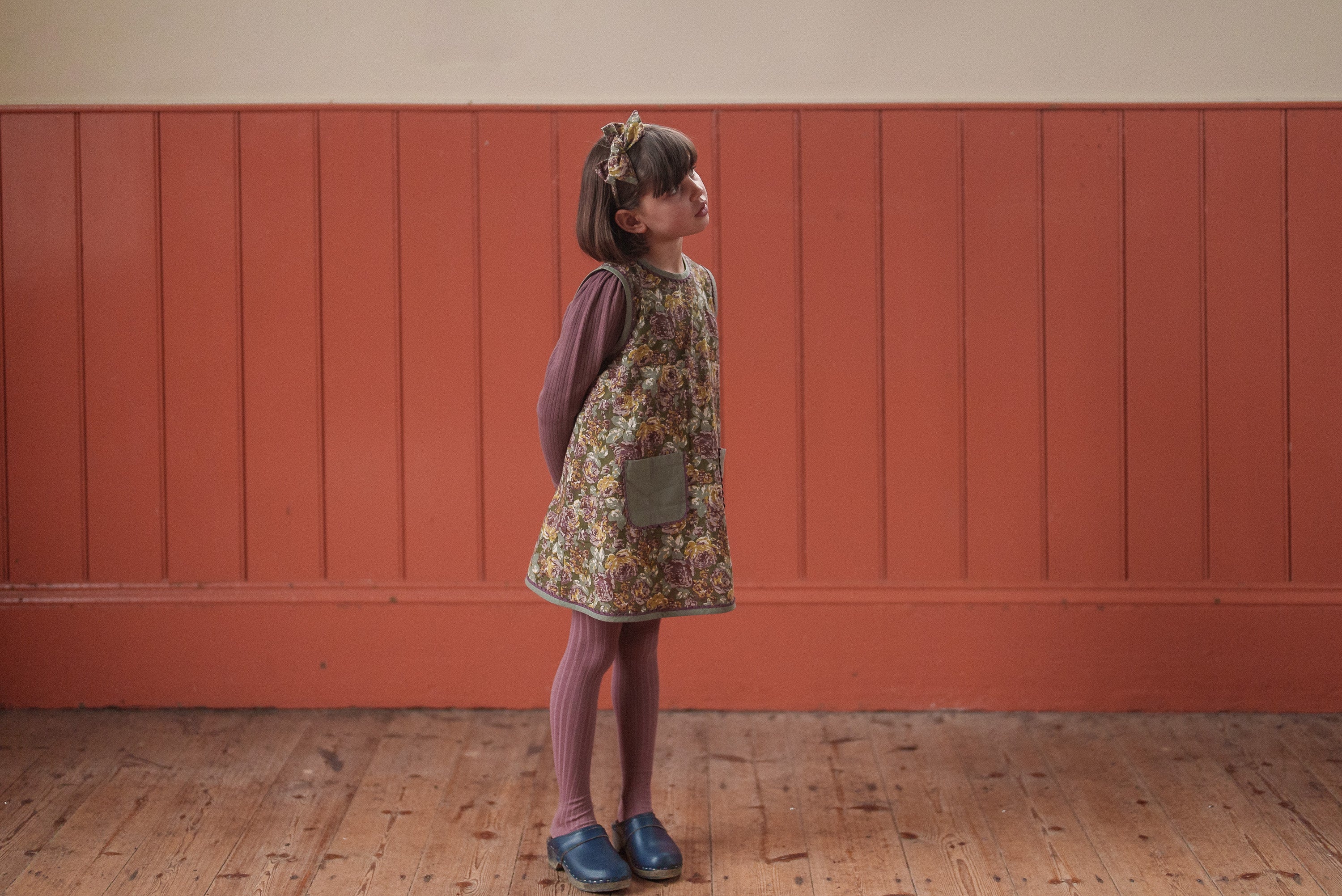 Young girl wearing a vintage style green floral organic cotton dress with tights and navy shoes, standing indoors against a warm-toned wooden wall.