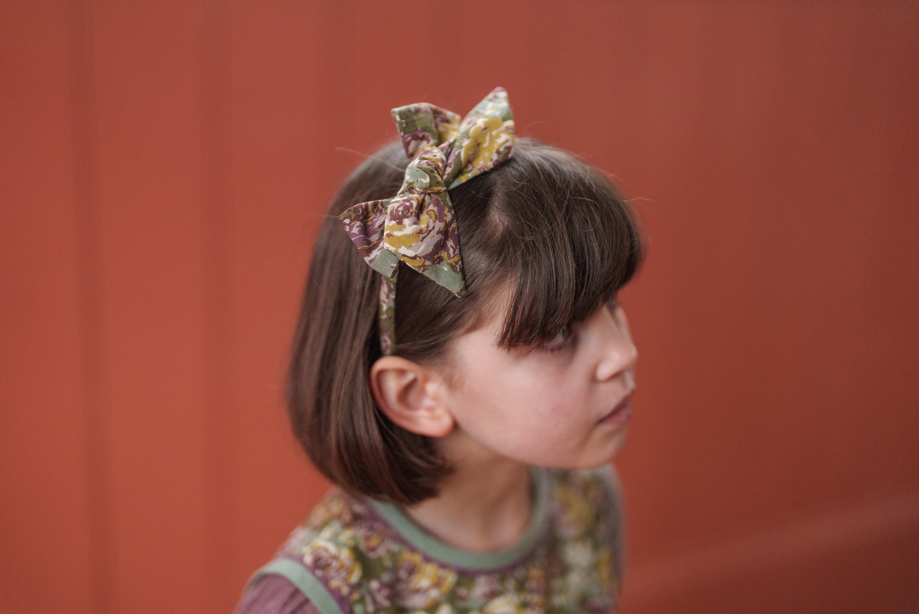 Young girl with a floral green dress made from organic cotton, wearing a matching vintage-style bow headband, showcasing sustainable and natural children's clothing.