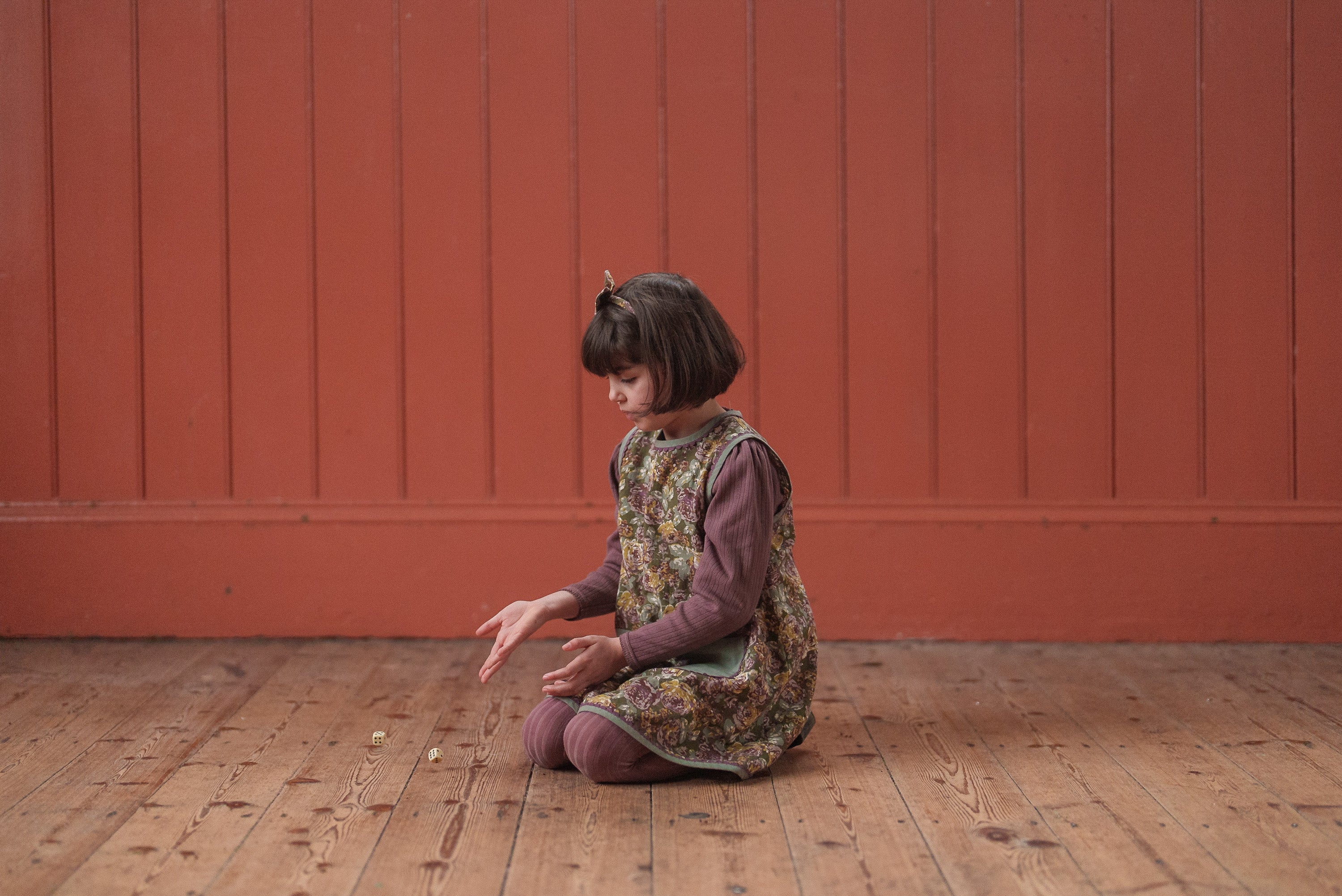 Young girl wearing a vintage style organic cotton floral dress in green, sitting on a wooden floor against a warm red wall, showcasing sustainable children's clothing.