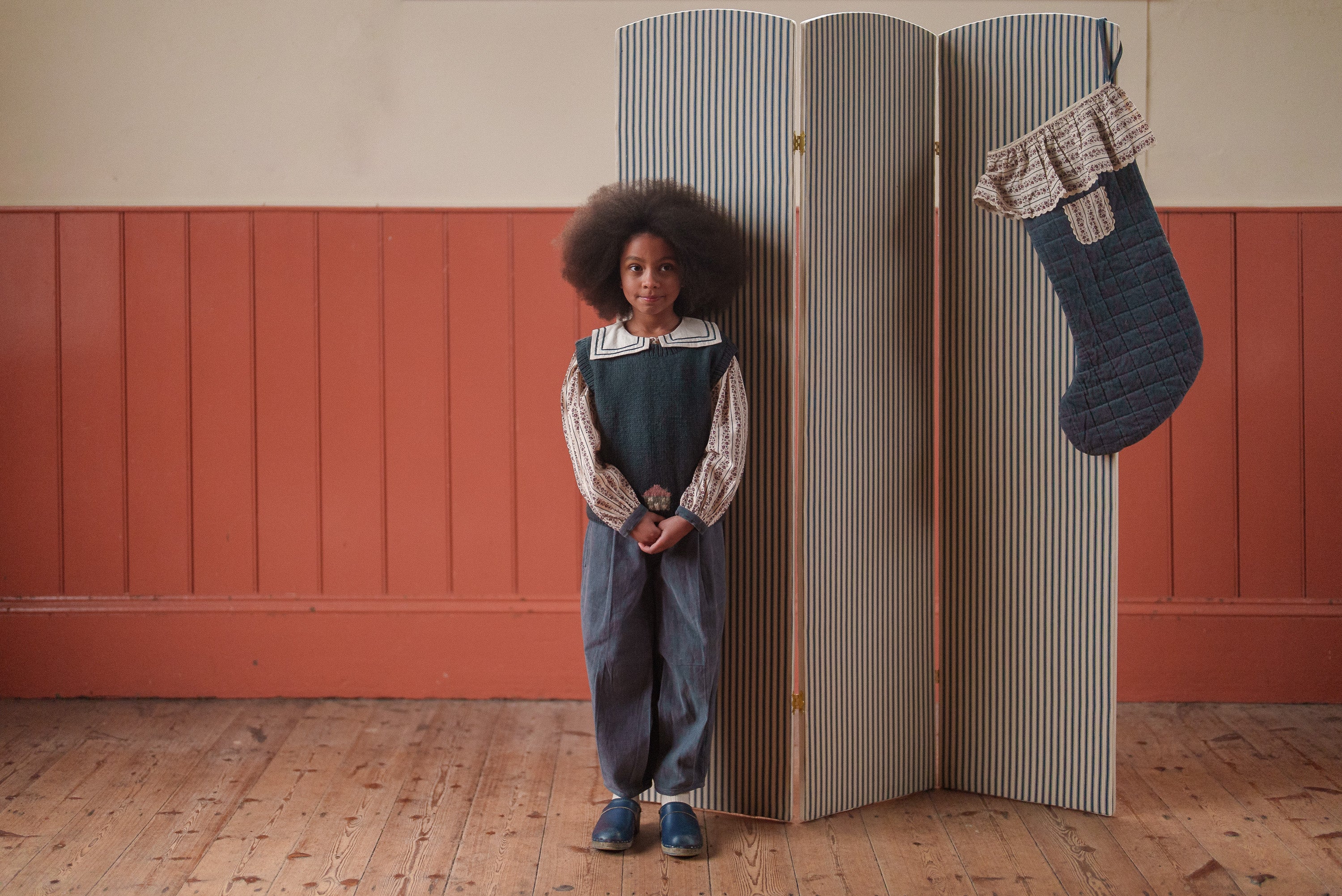 A young child wearing blue velvet trousers made from organic cotton, styled with a vintage-inspired top, standing in a softly lit room with warm wooden floors and a striped screen background.