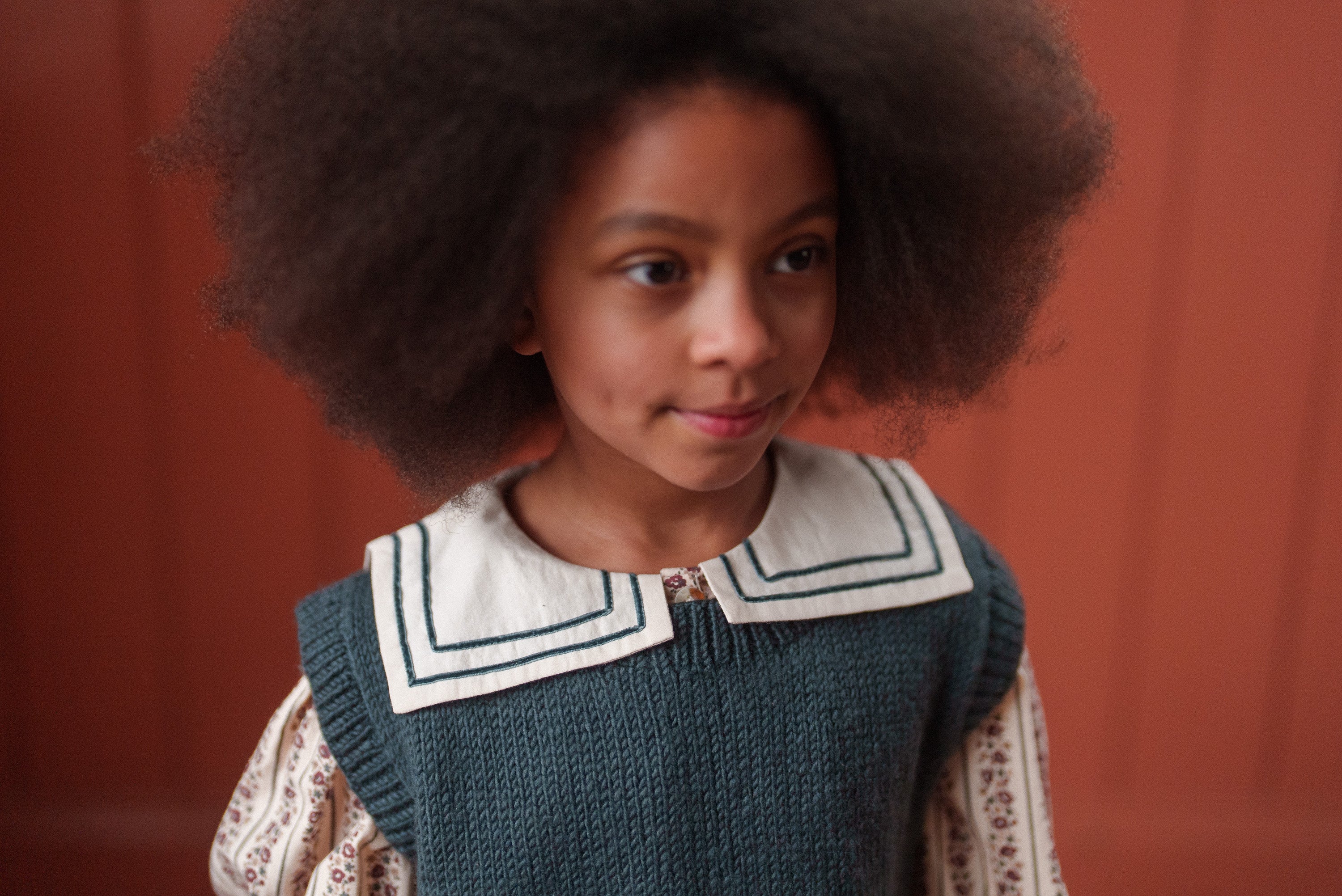 Young girl wearing a vintage style organic cotton blouse with a striped white collar and a blue knitted vest, showcasing ethically made sustainable children's clothing.