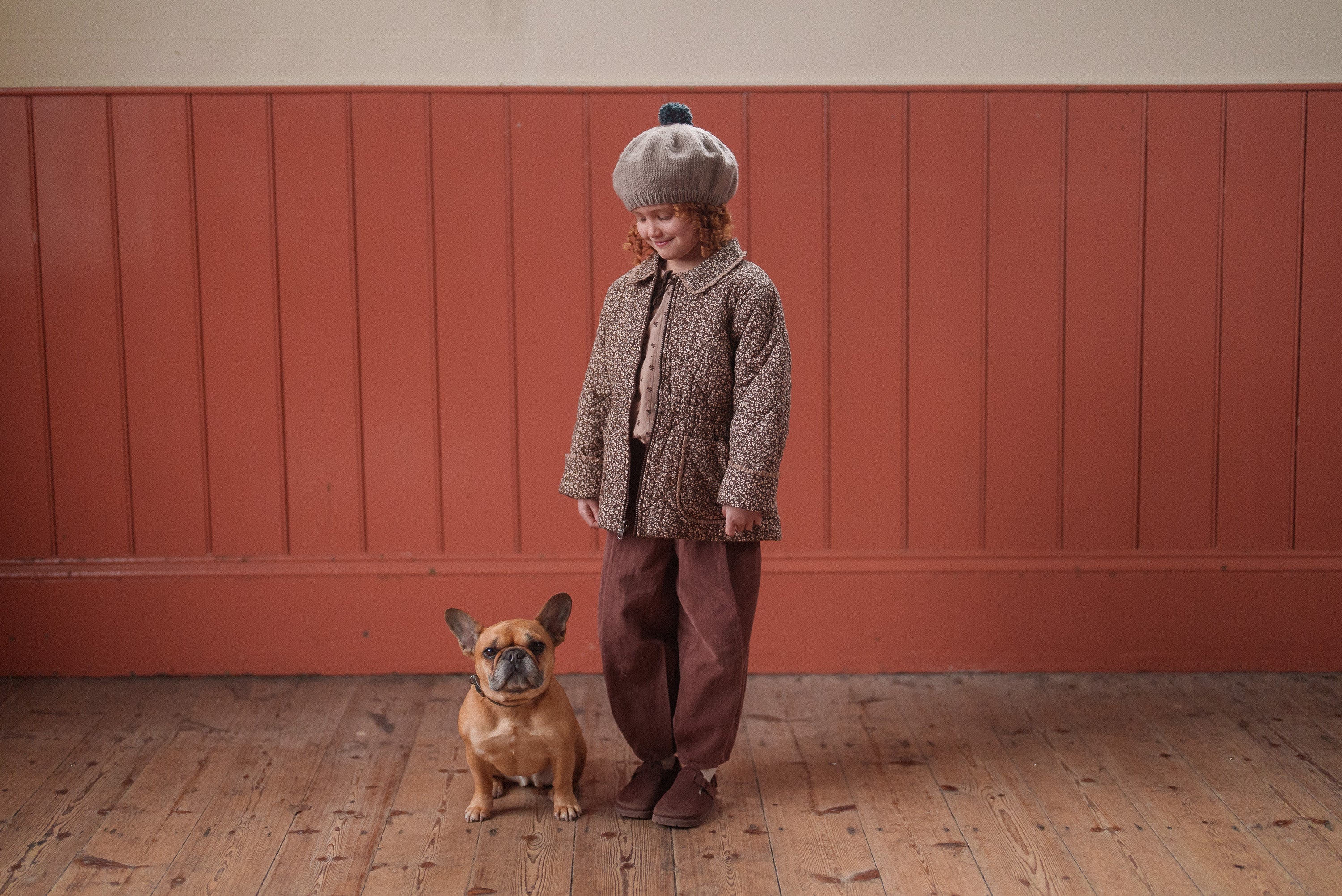 A child wearing a vintage-inspired brown and beige jacket made from organic cotton, paired with loose brown trousers and a wool hat, standing on a wooden floor next to a small dog.