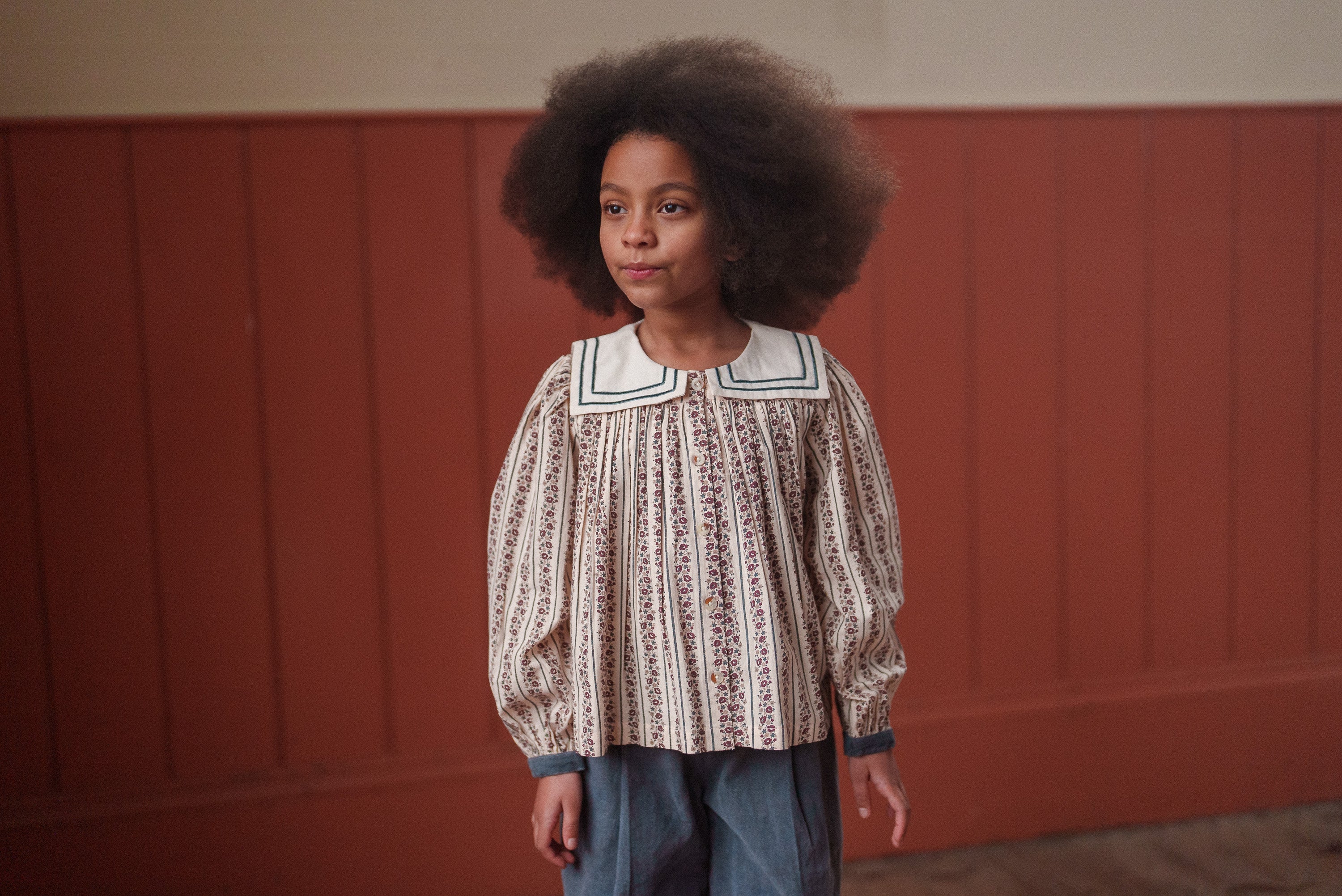 Young girl wearing a vintage style blouse made from organic cotton with a delicate floral pattern and traditional design, standing against a wooden panel background.