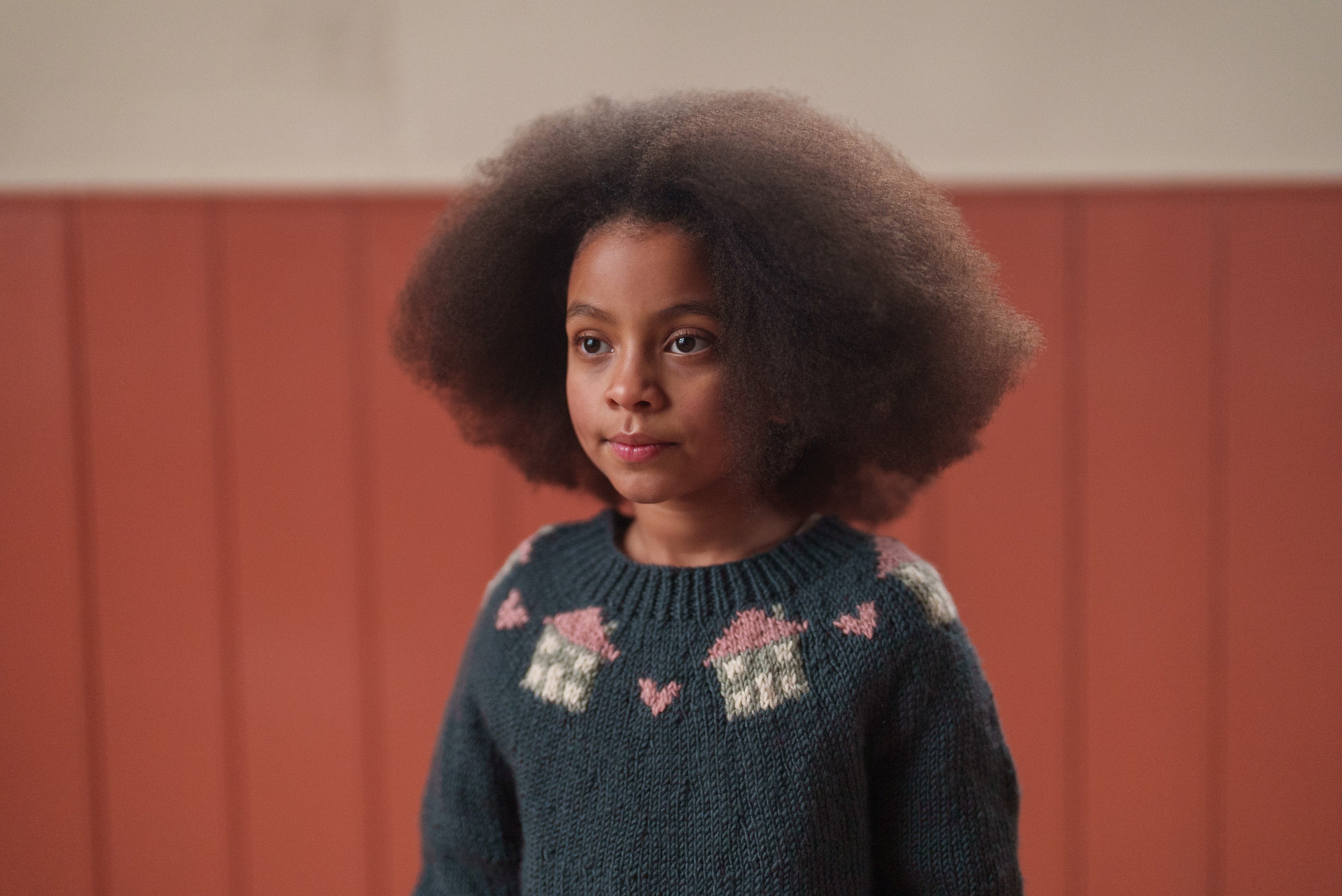 Young girl wearing a multicolor hand-knitted merino wool jumper with cottage motifs, showcasing slow, natural, ethical children’s clothing in a vintage style.