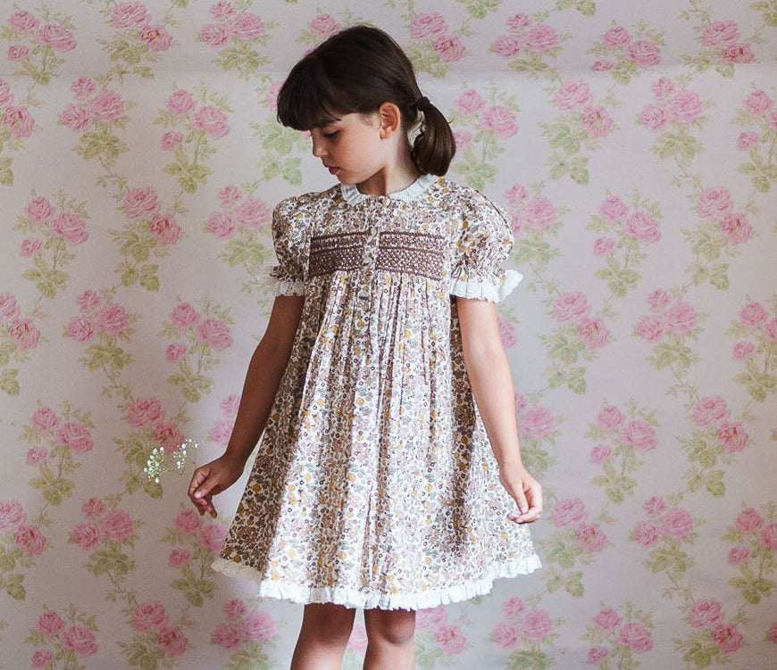 A young girl wearing a vintage style floral cotton blend dress with puff sleeves and smocked detailing, standing on a wooden floor against a floral wallpaper background.