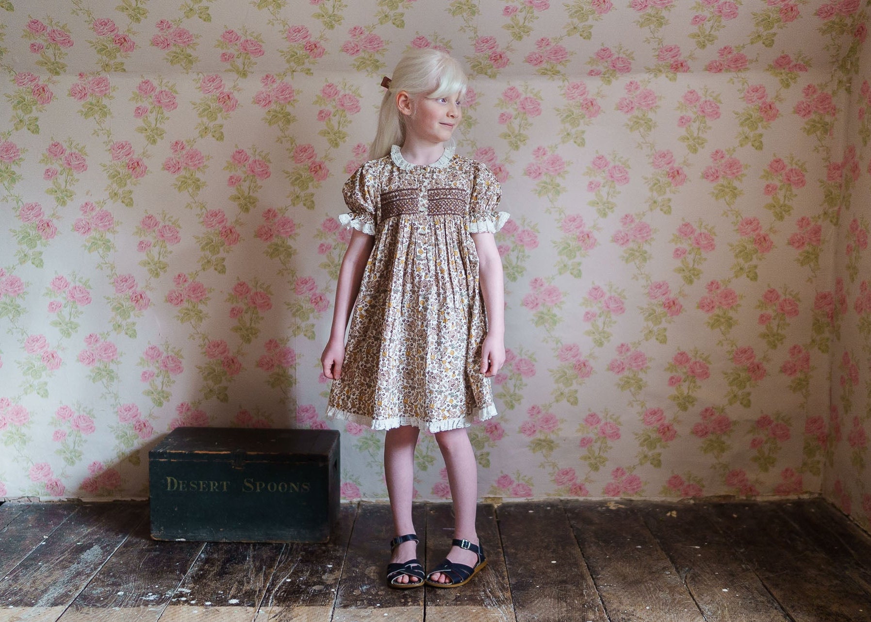 Young girl wearing a vintage-style floral cotton blend dress with smocked detailing, standing on rustic wooden floor against a floral wallpaper, evoking natural, ethical children’s clothing.