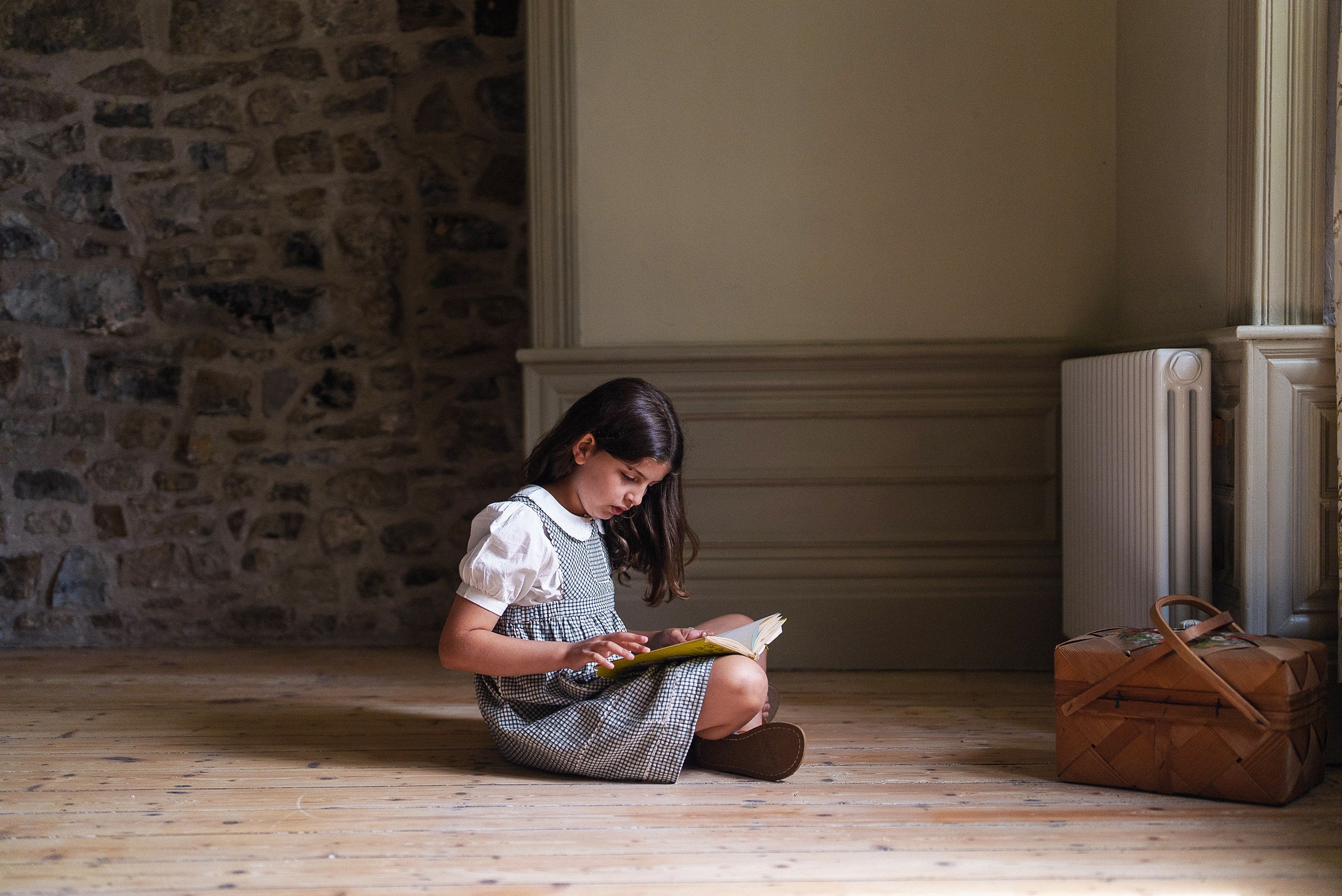 Young girl wearing an off-white organic cotton blouse with vintage style sitting on a wooden floor reading a book in a cozy, natural light room with stone wall background.