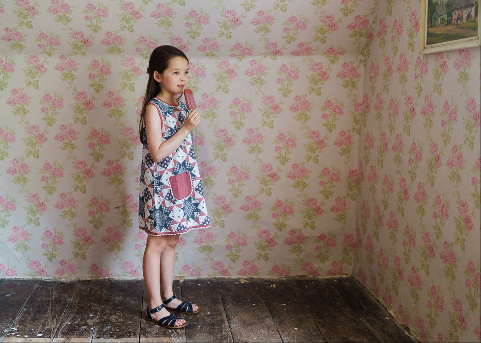 Young girl wearing a sustainable cotton blend floral patchwork dress, enjoying a treat in a vintage-style room with soft floral wallpaper, embodying natural, ethical children's clothing.