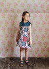 Young girl wearing a vintage-style cotton blend dress with a floral patchwork pattern and a scalloped collar, standing against a soft floral wallpaper background.
