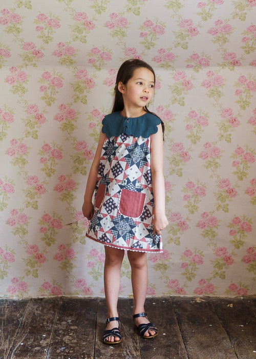 Young girl wearing a vintage-style cotton blend dress with a floral patchwork pattern and a scalloped collar, standing against a soft floral wallpaper background.