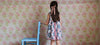 Young girl wearing a vintage style floral patchwork cotton dress with a pink ribbon in her hair, standing next to a blue wooden chair in a softly patterned room.