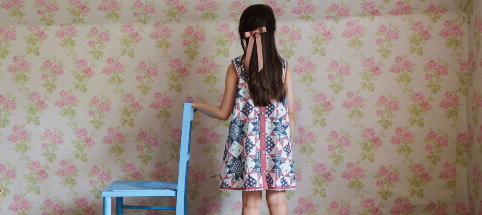 Young girl wearing a vintage style floral patchwork cotton dress with a pink ribbon in her hair, standing next to a blue wooden chair in a softly patterned room.