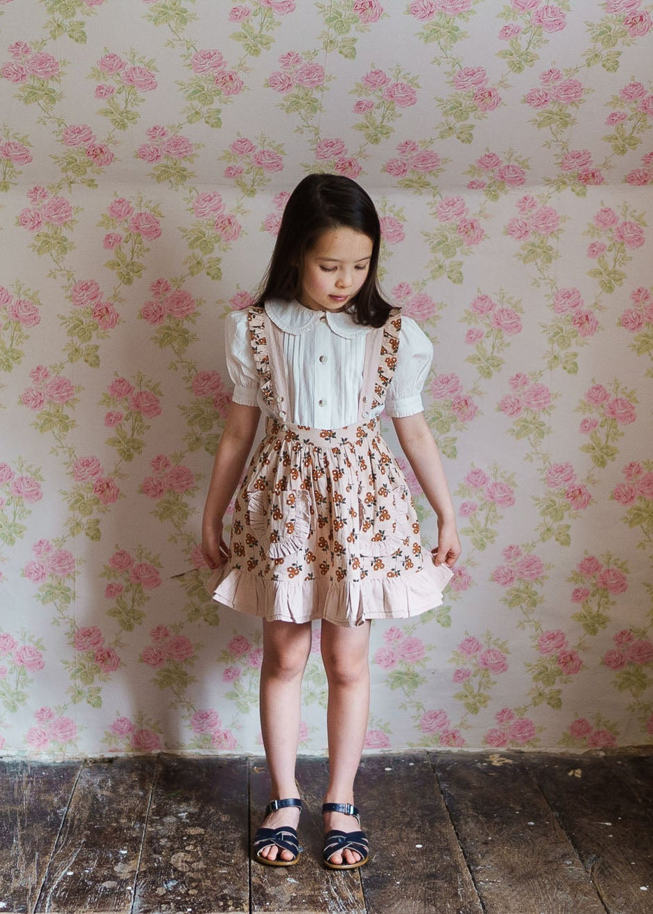 Young girl wearing vintage style floral organic cotton skirt with suspenders and a white blouse, standing on rustic wooden floor against floral wallpaper background.