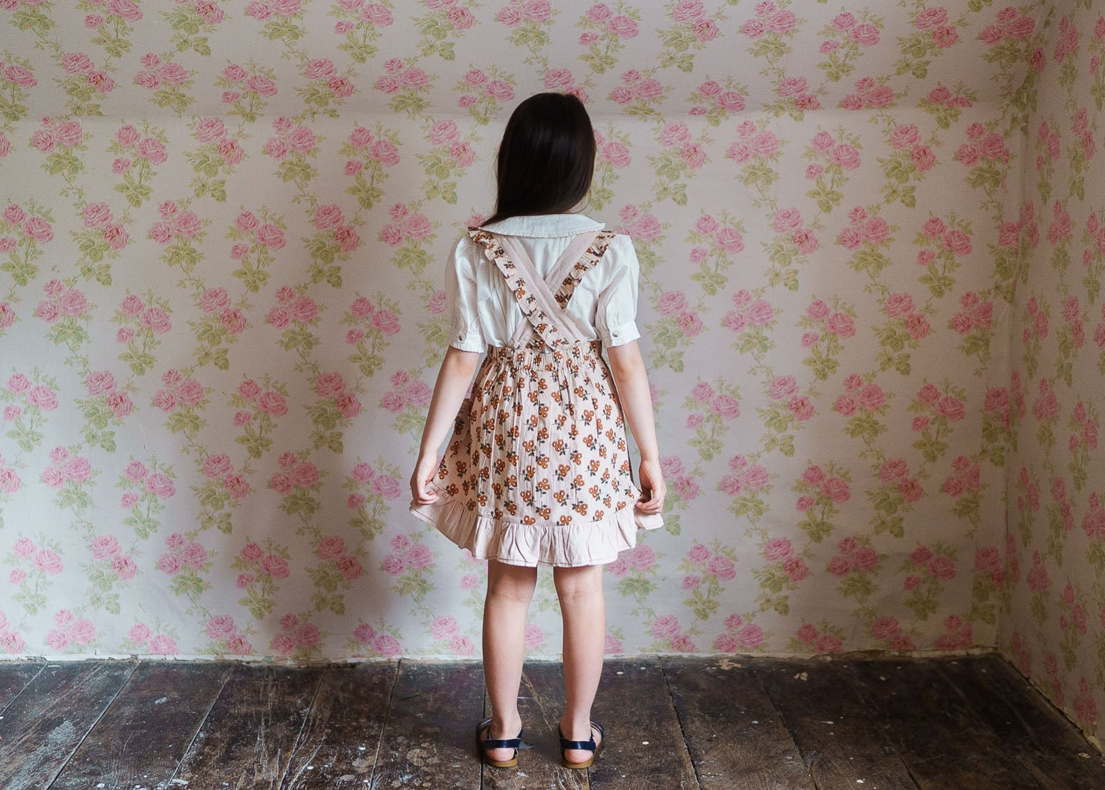Young girl wearing vintage style floral skirt with suspenders made from organic cotton, standing in a room with matching floral wallpaper, showcasing sustainable children’s clothing.