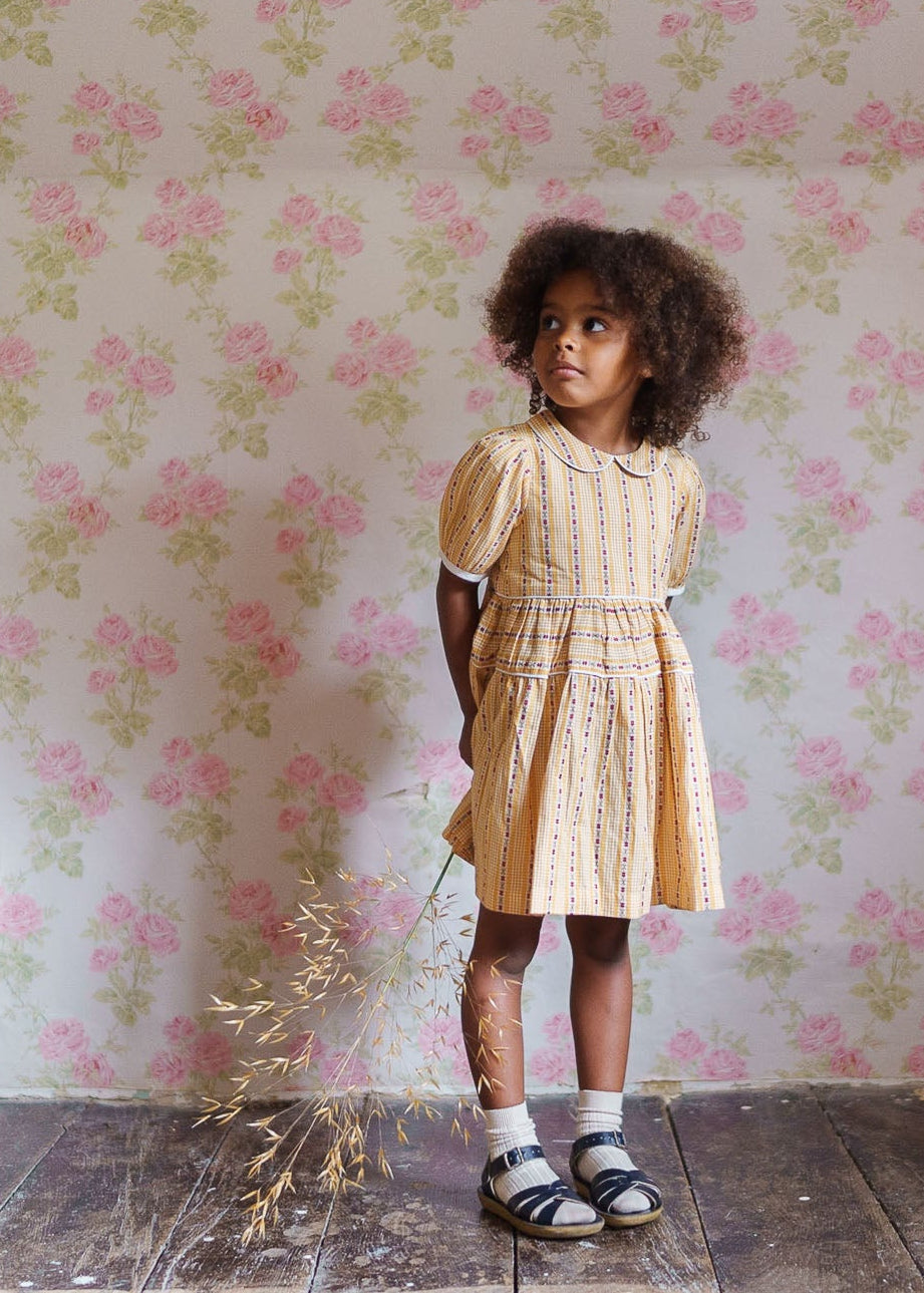 Young girl wearing a vintage-style yellow floral organic cotton dress with puffed sleeves and a Peter Pan collar, standing against pink floral wallpaper in natural light.