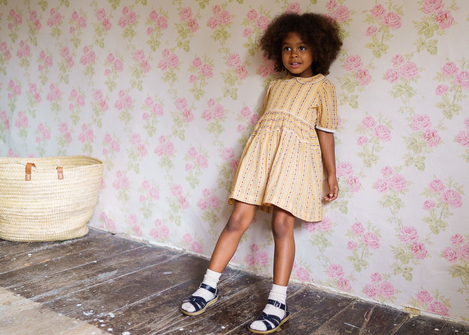 A young girl wearing a yellow floral dress made of organic cotton, standing by a vintage-style floral wallpaper in a sustainably made, ethically designed children's outfit.