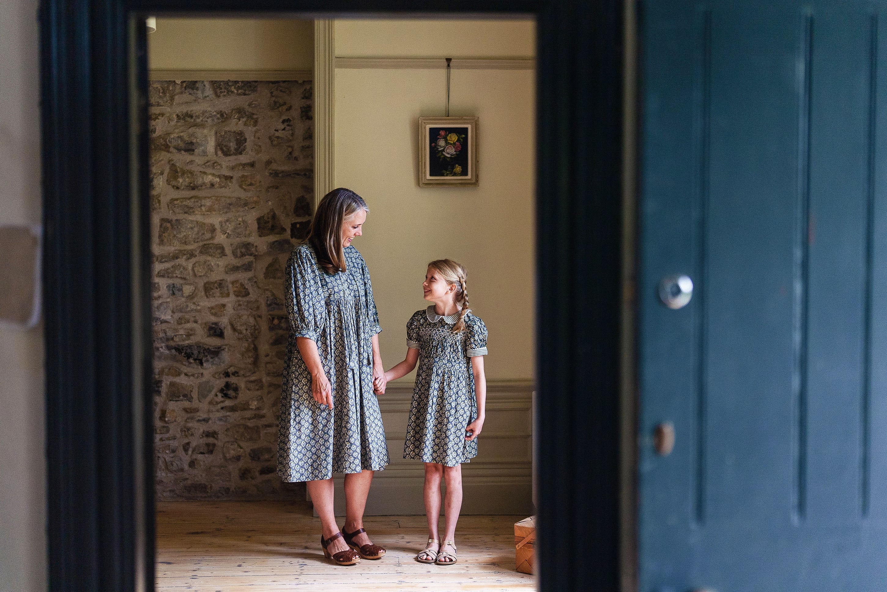 A woman and a young girl wearing matching floral cotton dresses stand holding hands in a cozy vintage-style room, showcasing sustainable, ethically made clothing.