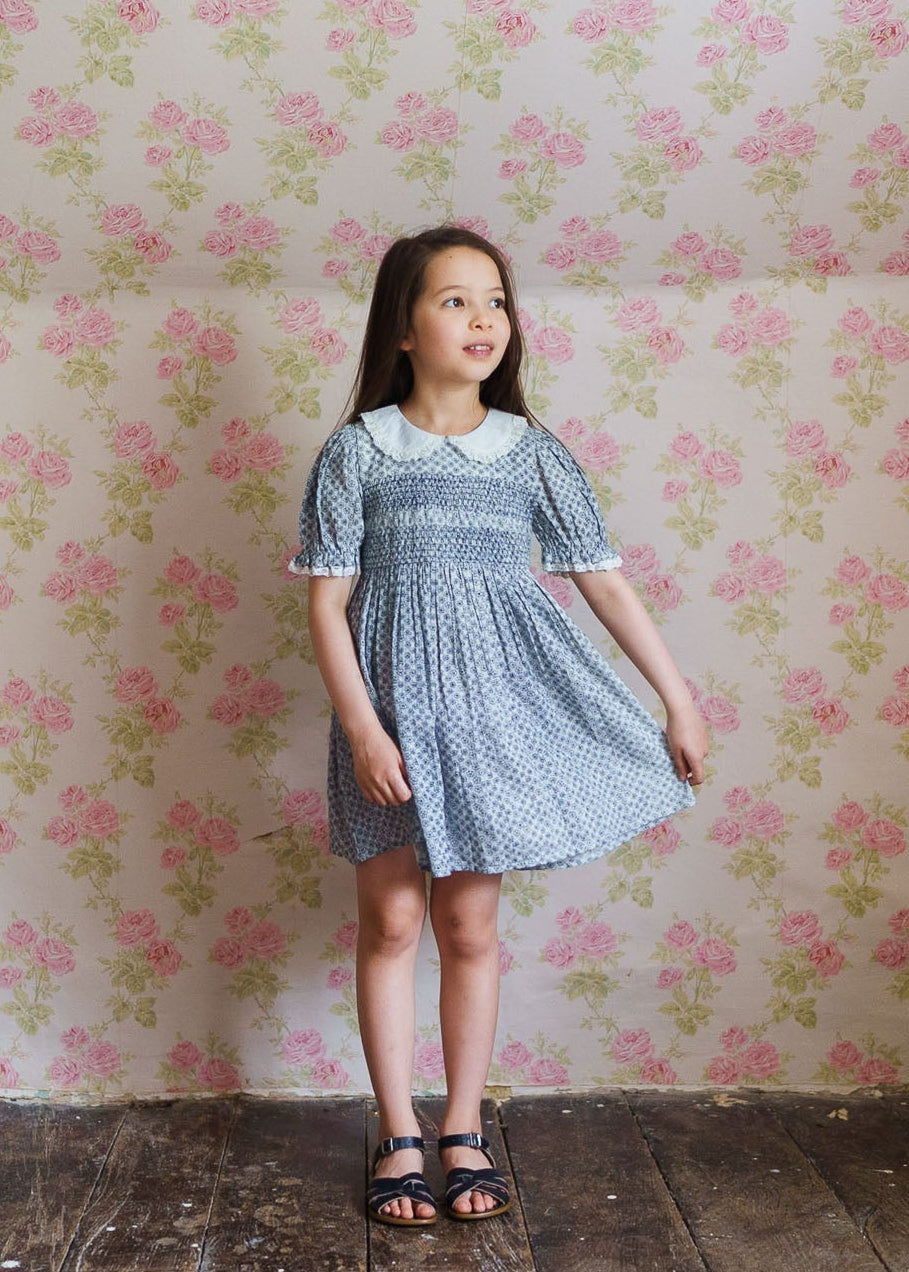 Young girl wearing a vintage style blue floral smocked dress made from a cotton blend, standing against a pink rose patterned wallpaper, showcasing natural, sustainable children's clothing.