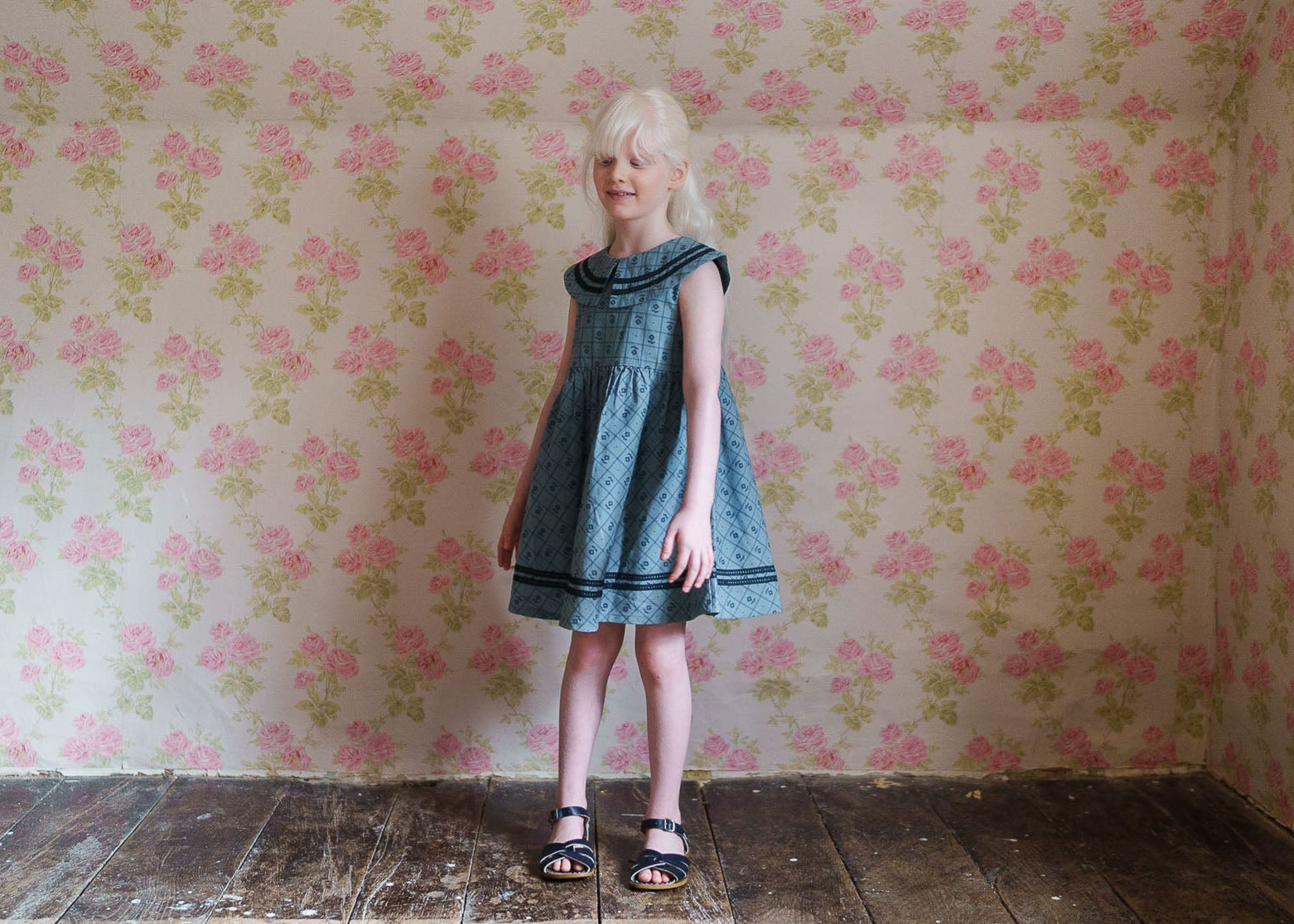Young girl wearing a vintage style girls dress in organic cotton featuring a delicate floral pattern, standing against a softly floral wallpaper background.