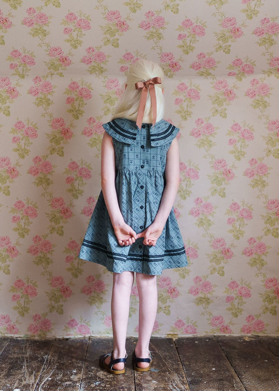 Young girl with long blonde hair tied with a ribbon wearing a vintage style organic cotton dress with a floral pattern, standing against floral wallpaper in a rustic room.