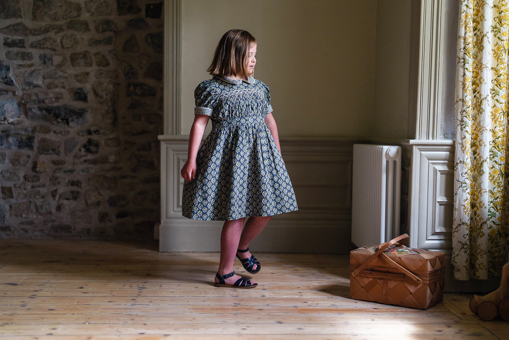Our timeless, vintage inspired Orla Dress has been made in our organic cotton thistle floral fabric. This pretty children's dress is perfect for special occasions or everyday wear. It has beautiful hand smocking over a gathered bodice, full, puffed sleeves with contrasting cuffs and a contrasting Peter Pan collar. Designed for comfort and style, the full skirt allows for easy movement, it's sure to become a favourite for little girls.  Little Girl holdi ng skirt and twirling. 
