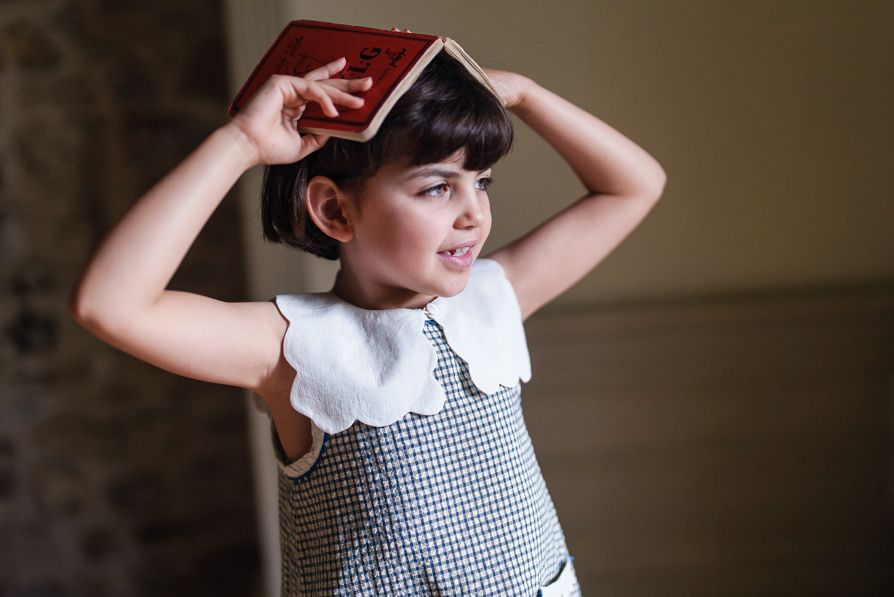 Young girl wearing vintage-style kids clothing with a white scalloped collar made from organic cotton, holding a book above her head in a warm, natural setting.