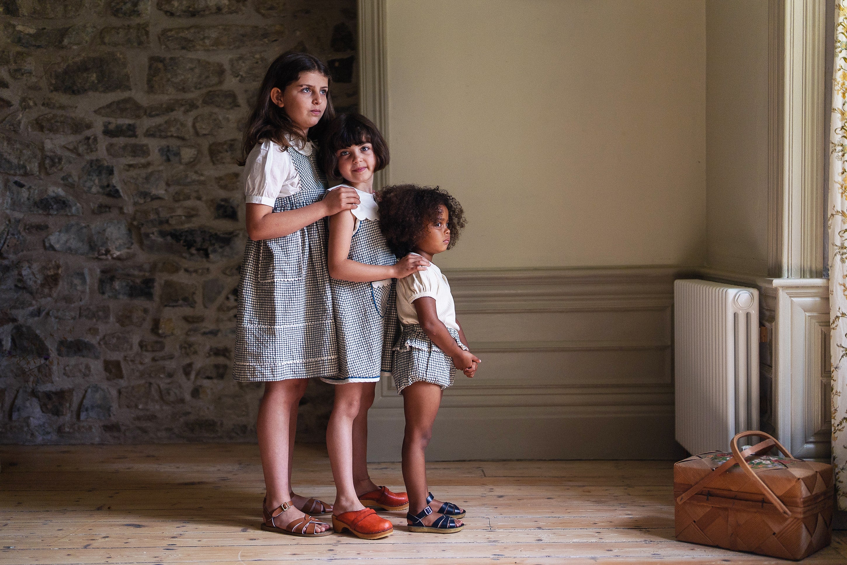 Three children wearing vintage style children's clothing made from organic cotton stand indoors by a stone wall and window, showcasing sustainable and ethically made kids clothing in natural tones.
