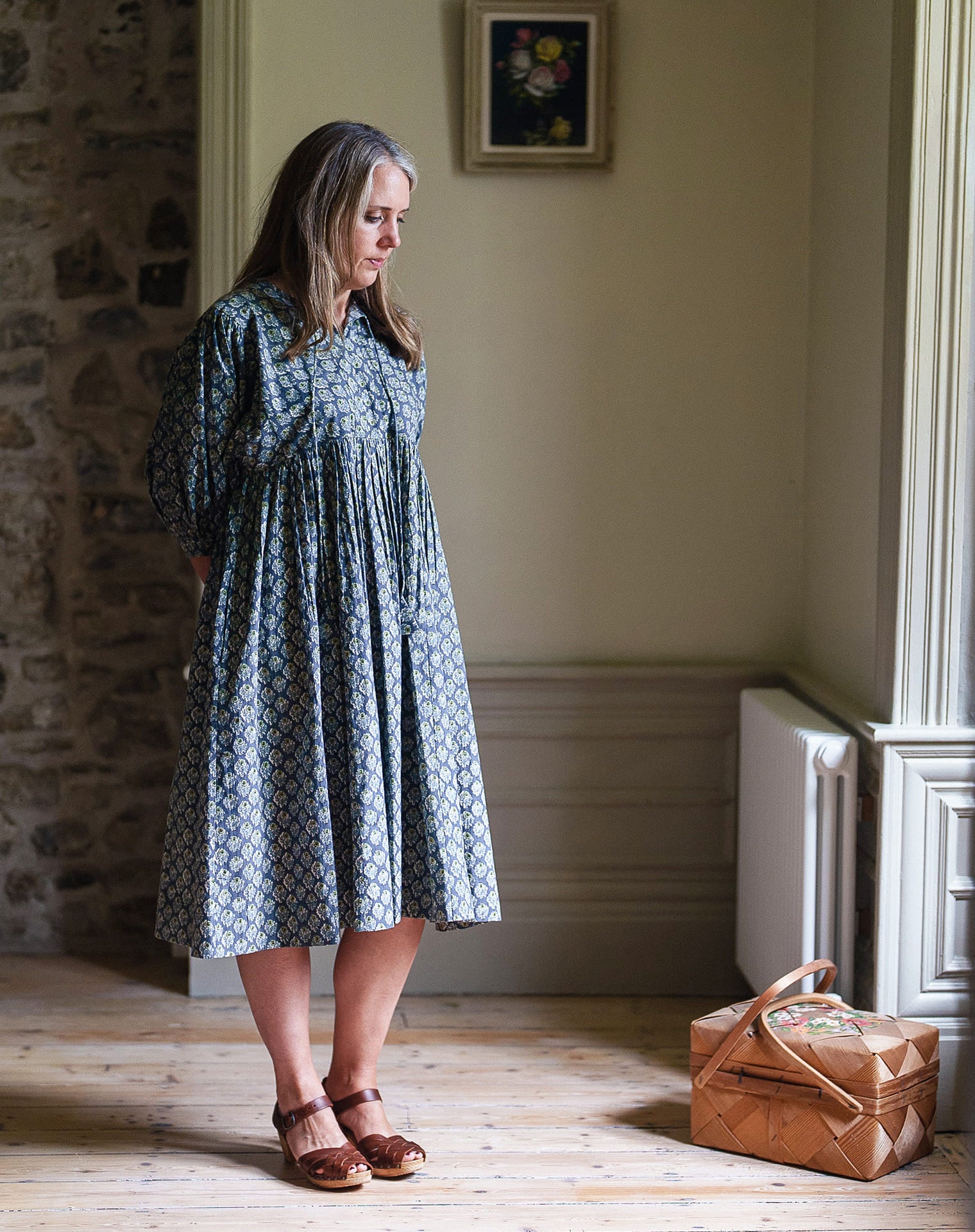 Woman wearing a vintage-style cotton dress with a subtle floral pattern, standing in a naturally lit room with wooden floors and a woven basket nearby, embodying slow, ethical fashion.