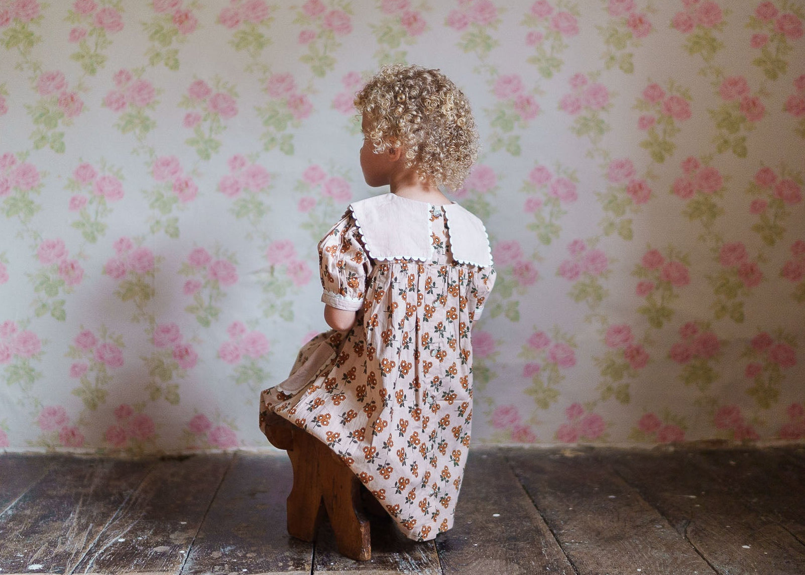 A young child with curly hair wearing a vintage-style organic cotton dress with a floral pattern, sitting on a wooden stool against a floral wallpaper background.