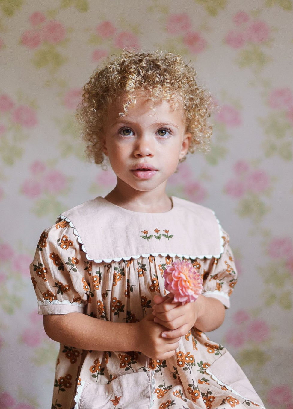 Young child wearing a vintage-style girls dress in floral organic cotton, holding a pink flower against a soft floral wallpaper background, showcasing natural and sustainable kids clothing.