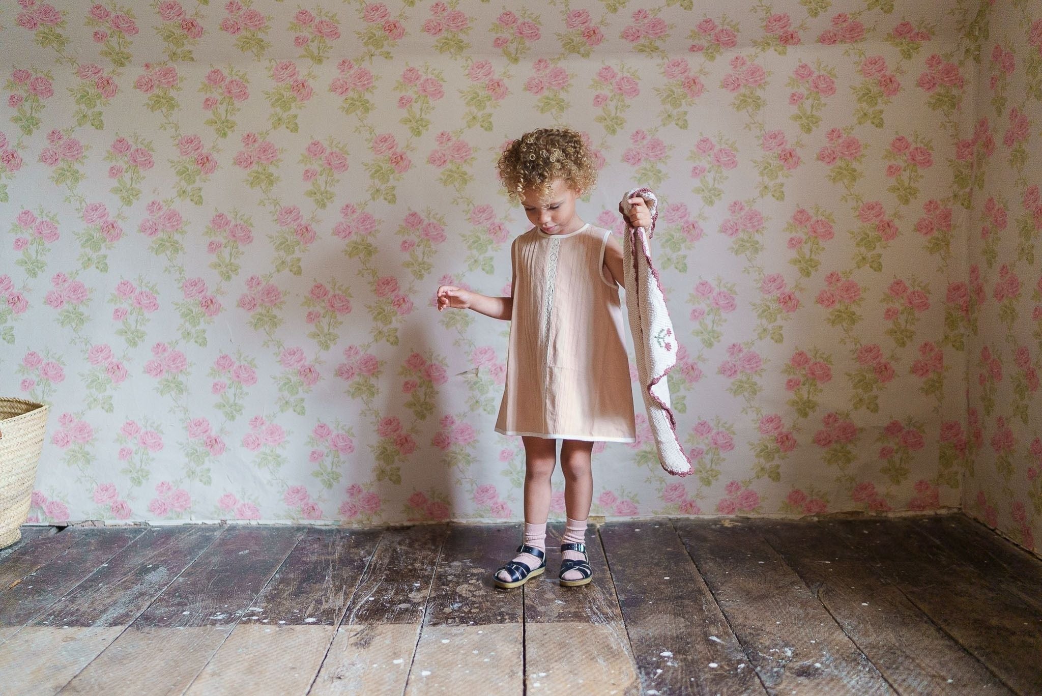 Young child wearing a powder pink organic cotton dress standing on a rustic wooden floor against floral vintage wallpaper, embodying natural and ethical children's clothing.
