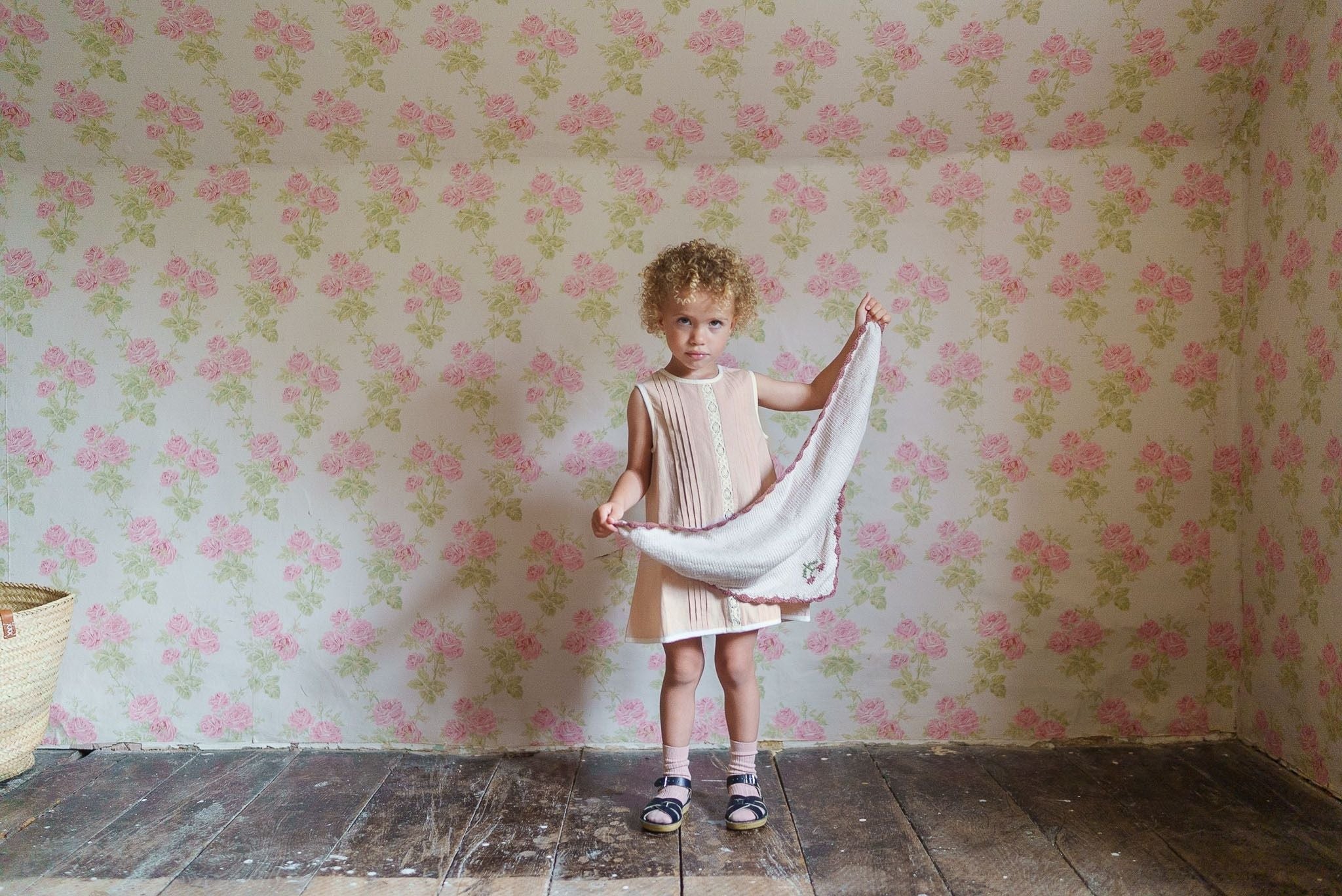 Young girl wearing a powder pink organic cotton dress with vintage style, standing on wooden floor against floral wallpaper, holding a white cloth.