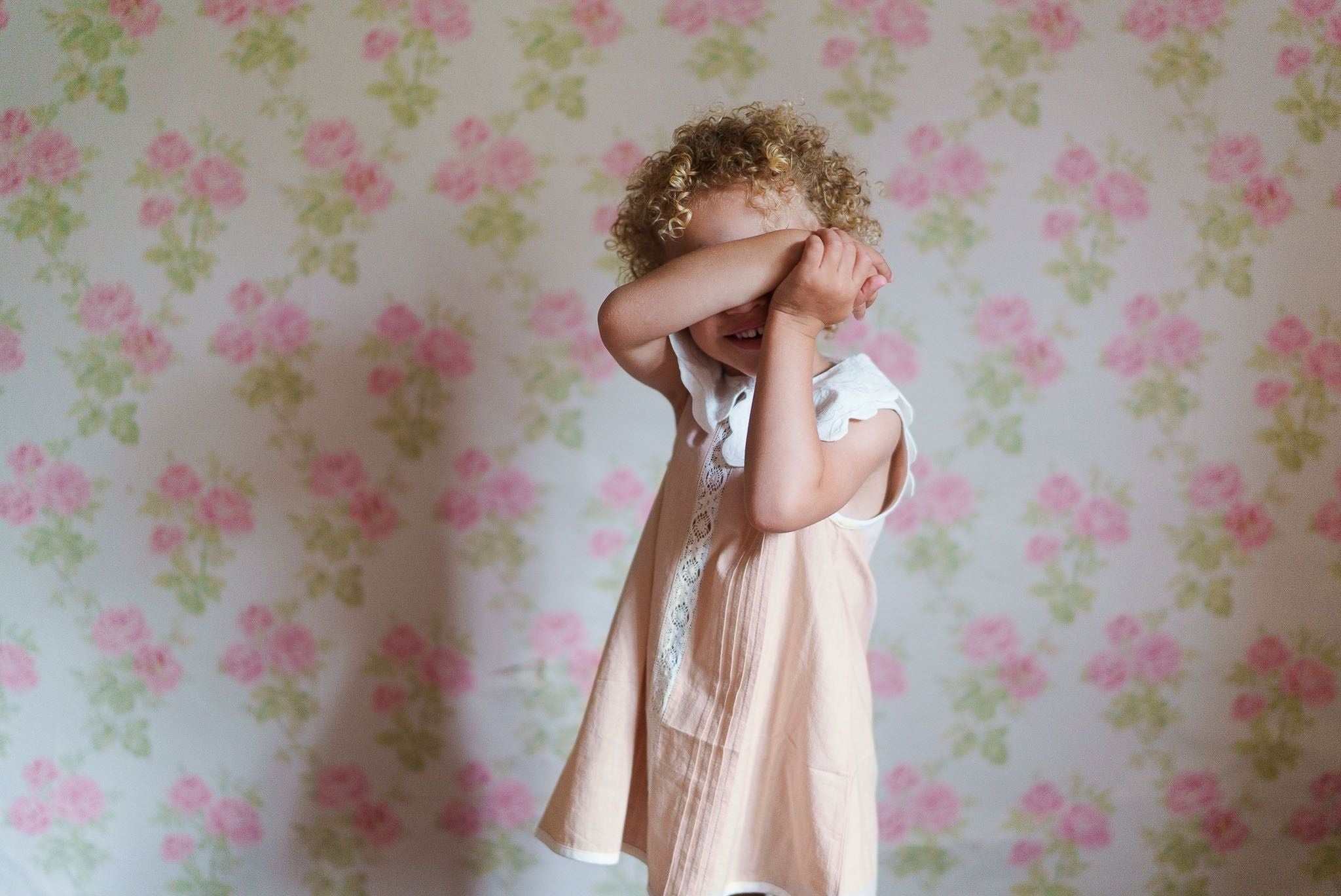 Young girl wearing a powder pink organic cotton dress with vintage style details, standing in front of a floral wallpaper background, embracing natural children's clothing.