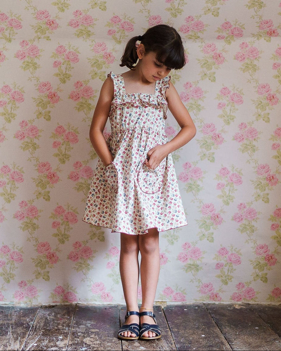 Young girl wearing a vintage style organic cotton dress with a delicate floral pattern, standing against a coordinating floral wallpaper in a warm, natural setting.