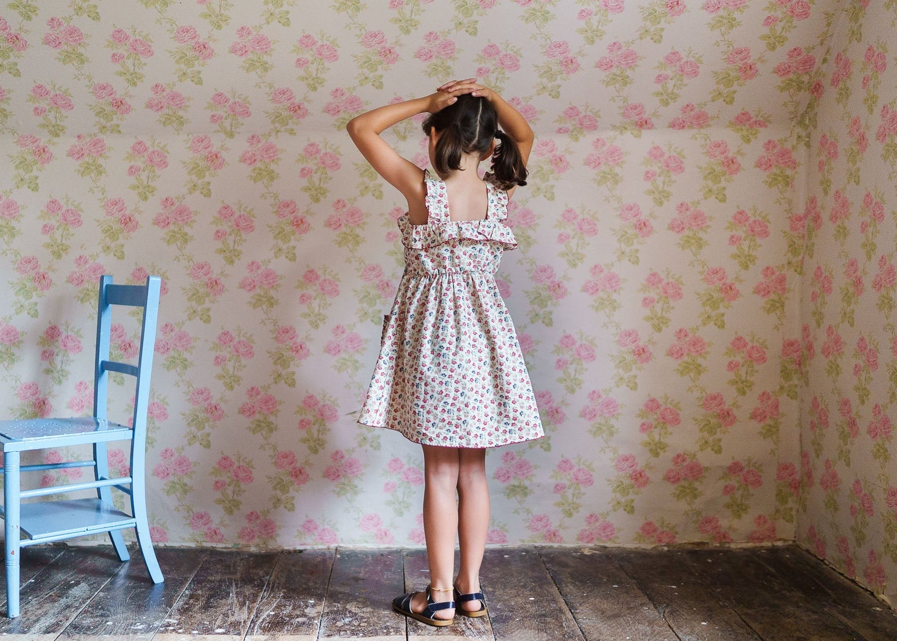Young girl wearing a vintage-style floral organic cotton dress in a softly patterned room with rustic wooden floors and a pastel blue chair nearby.