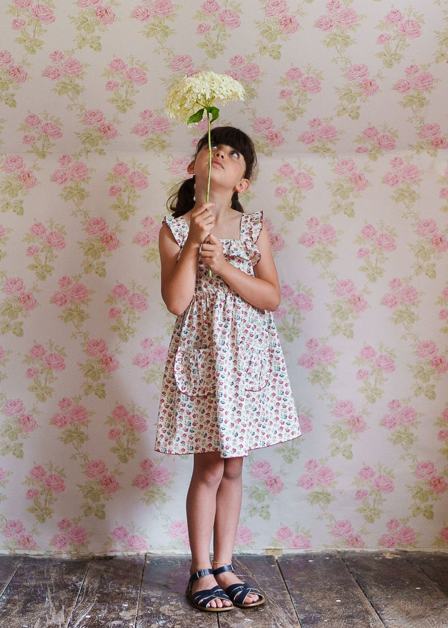 Young girl wearing a sustainable organic cotton floral dress with ruffle details, standing on a wooden floor against vintage floral wallpaper, holding a white flower.