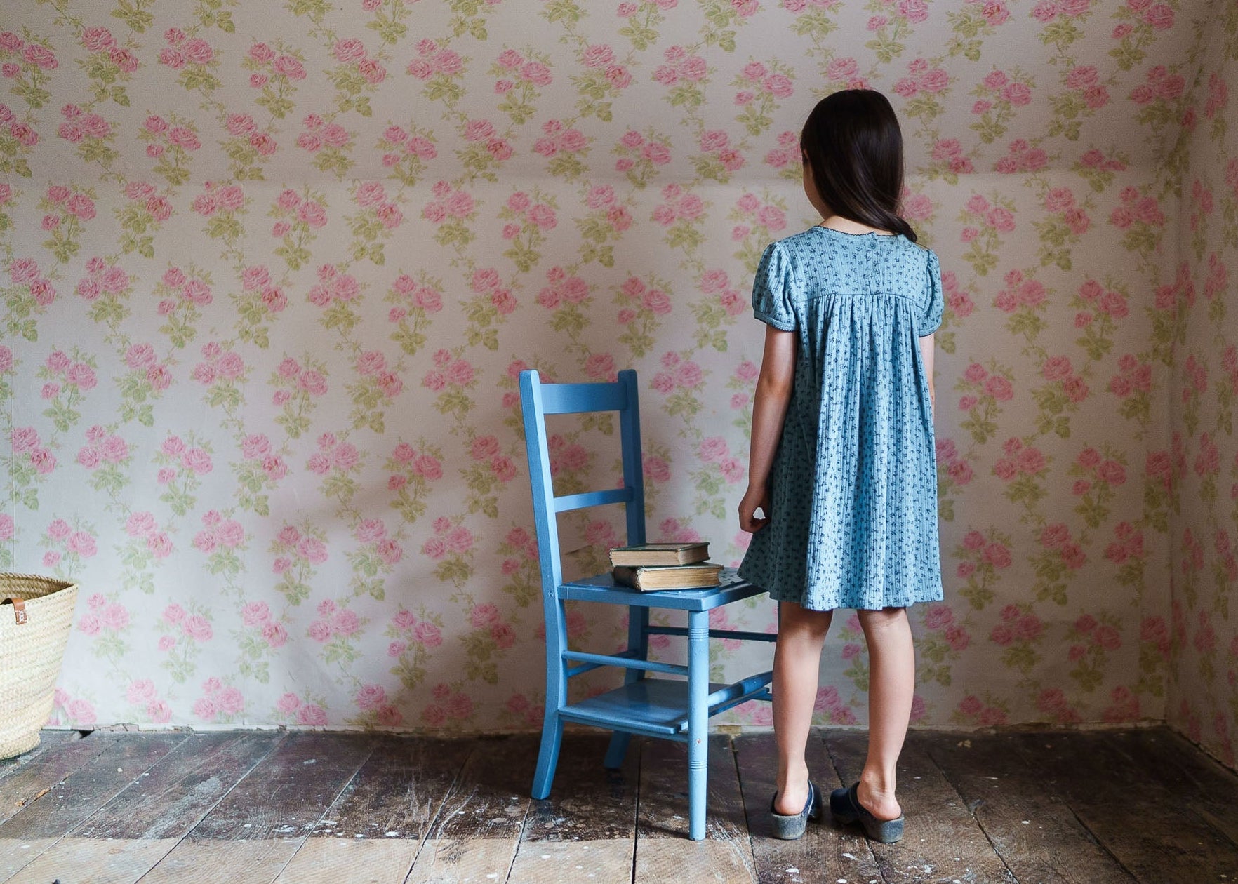 Young girl wearing a blue floral organic cotton dress standing by a vintage wooden chair with books, in a softly lit room with floral wallpaper and rustic wooden floor.