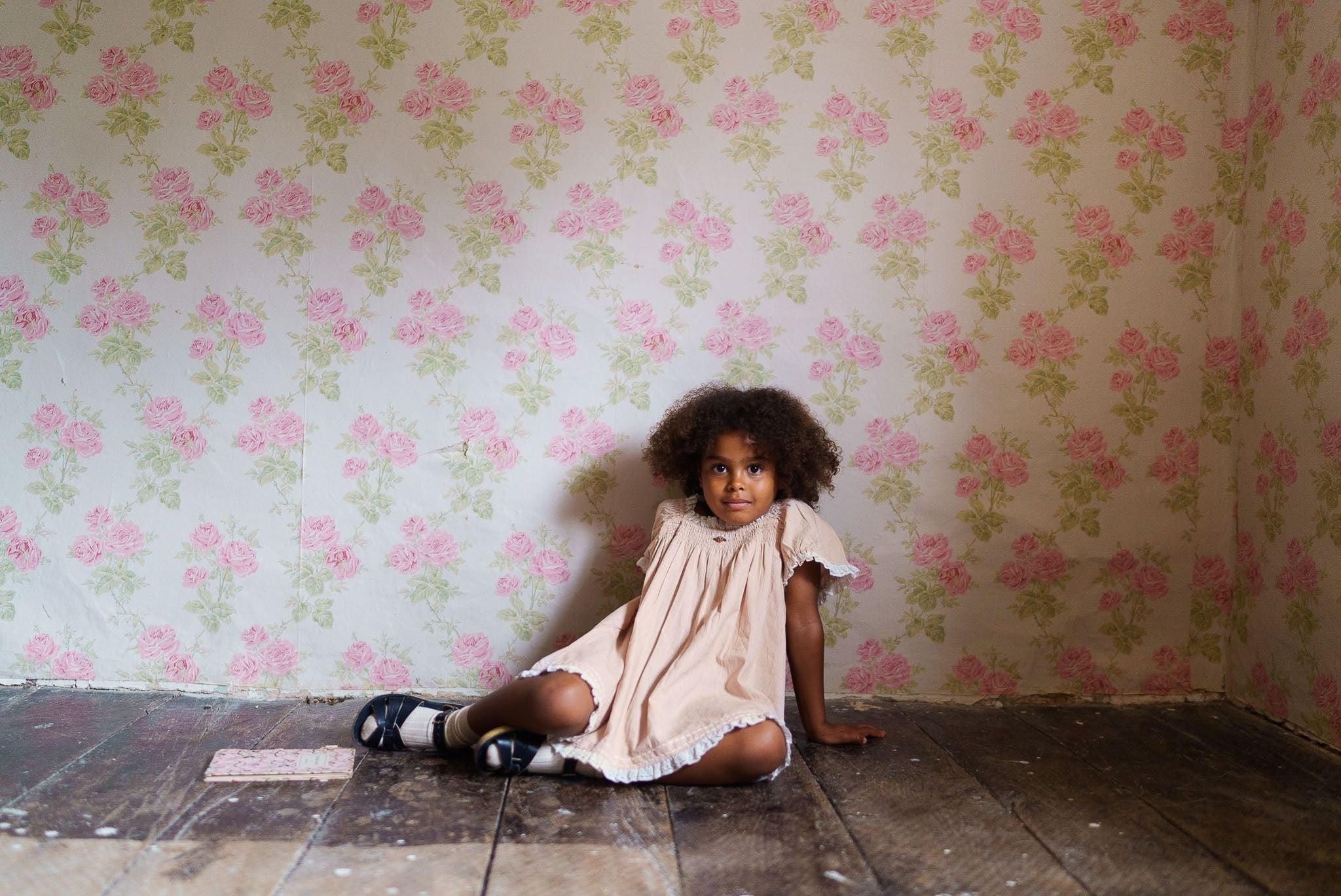 Young girl wearing a pink organic cotton dress with a vintage style, sitting on a wooden floor against a floral patterned wall in a natural, ethical children’s clothing setting.