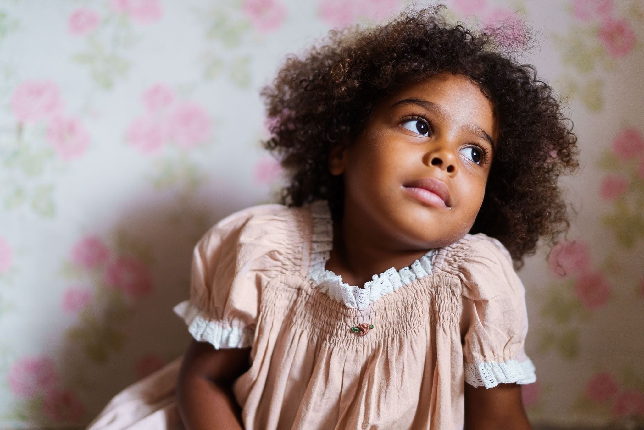 Young girl wearing a vintage-style pink organic cotton dress with delicate white lace trim, sitting thoughtfully against a floral wallpaper backdrop.
