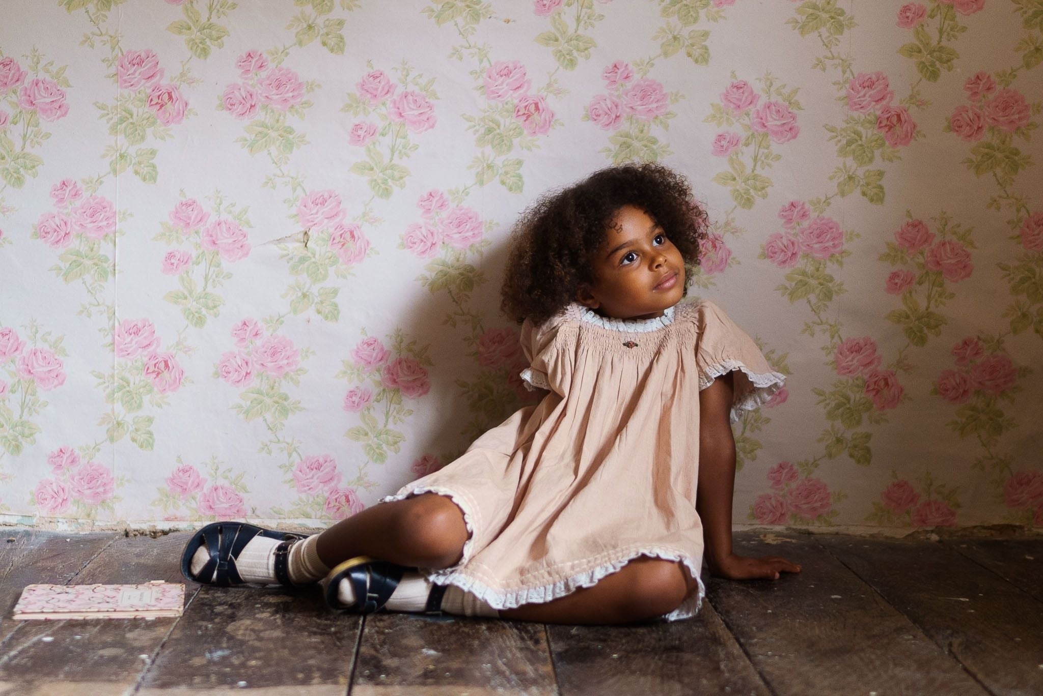 Young girl wearing a powder pink organic cotton vintage-style dress sitting on rustic wooden floor against a floral wallpaper, showcasing sustainable children's clothing.