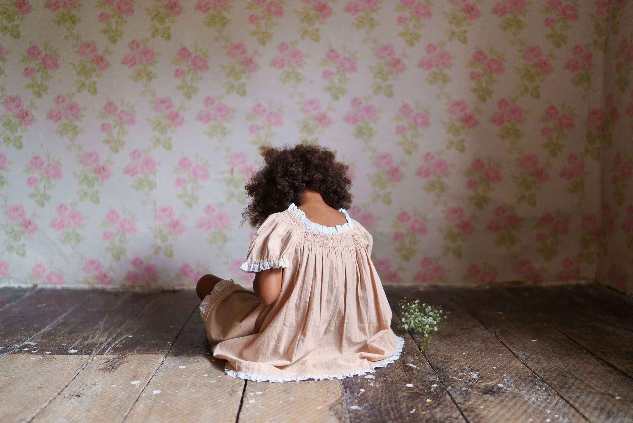 Young child sits on rustic wooden floor wearing a pink organic cotton vintage-style dress with delicate lace trim, against a floral wallpaper background.