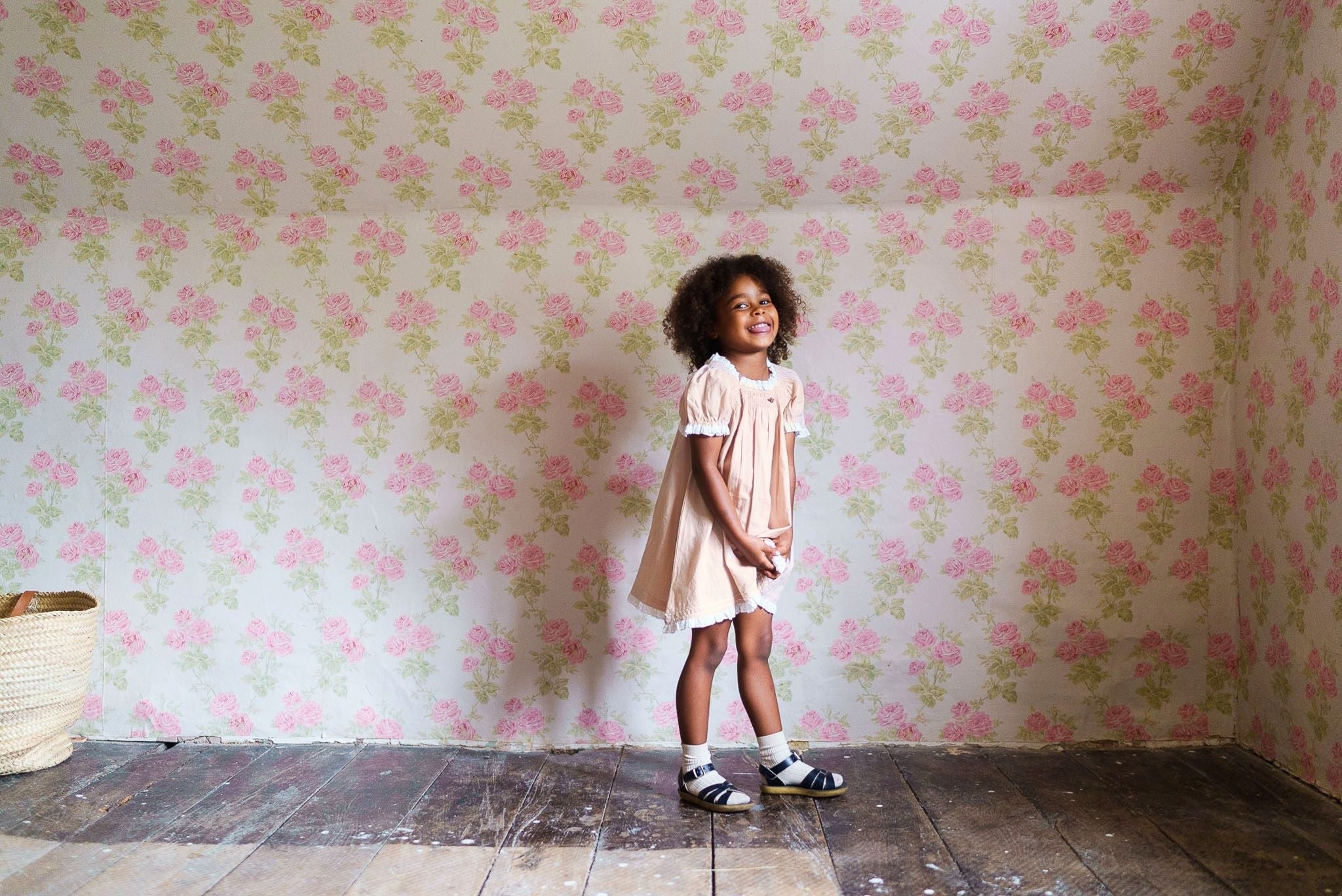 Young girl wearing a powder pink organic cotton vintage style dress standing in a bright room with floral wallpaper, showcasing sustainable children's clothing.