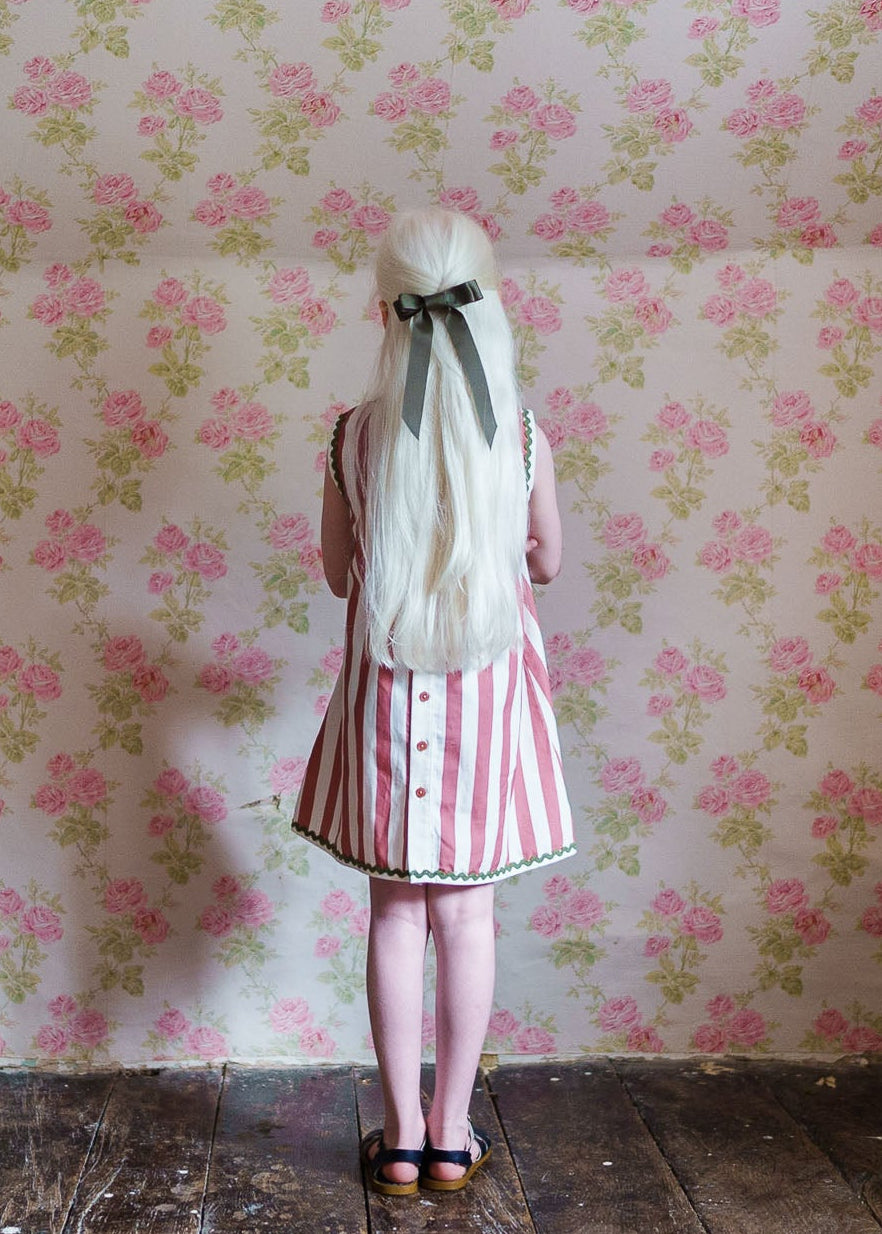 Young girl with long blonde hair tied with a black ribbon standing against floral vintage wallpaper, wearing a pink and white striped organic cotton dress with button details.